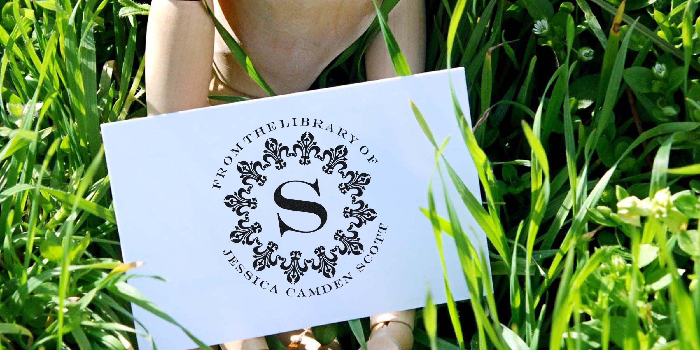 White card with an ornate circular emblem and a large letter S, reading “From the Library of Jessica Camden Scott,” placed on green grass.