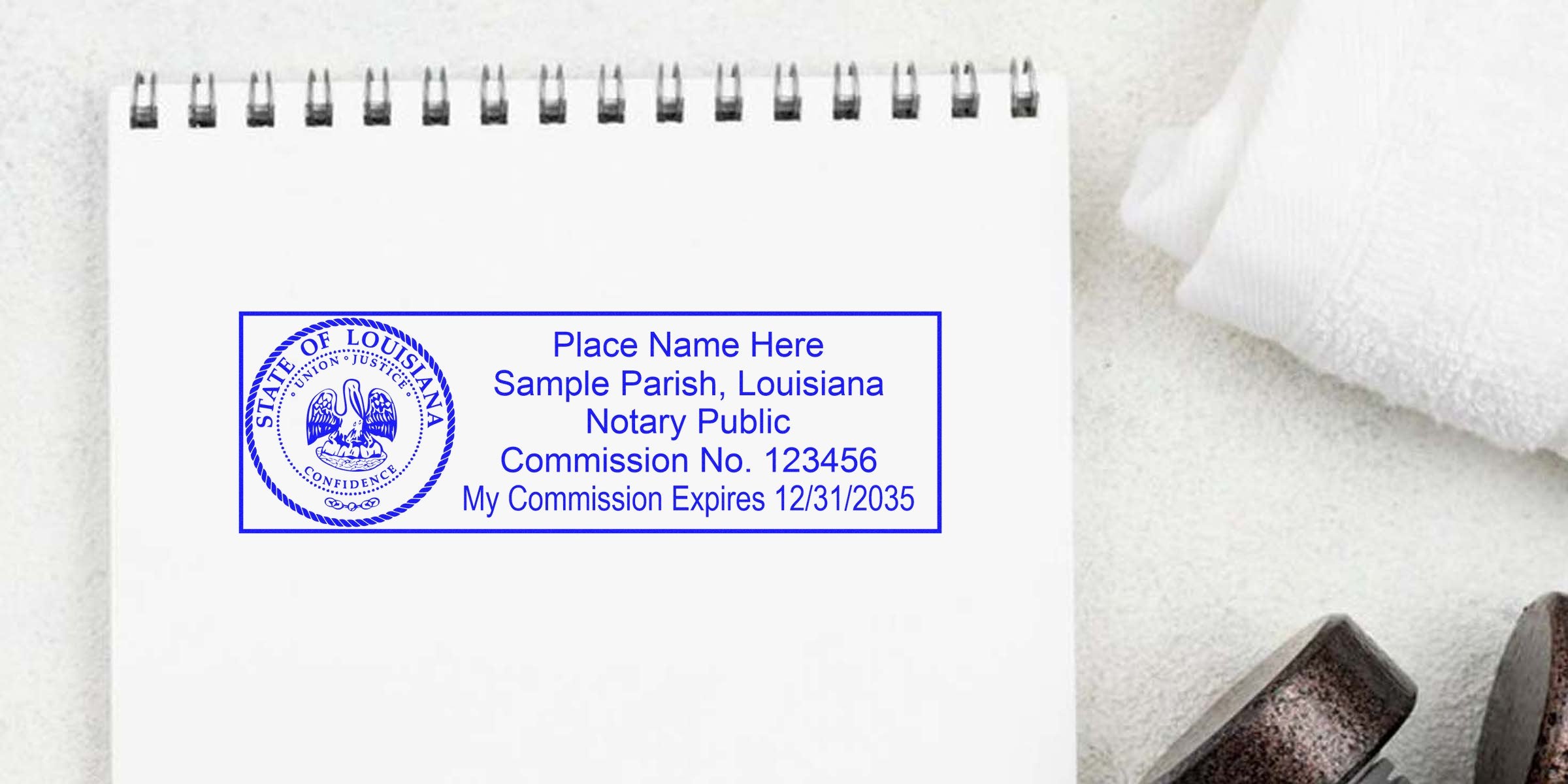 Blue Louisiana notary public stamp on white spiral notebook showing state seal, sample name, parish, commission number and expiration date, close-up with towel and dumbbell in soft textured background.