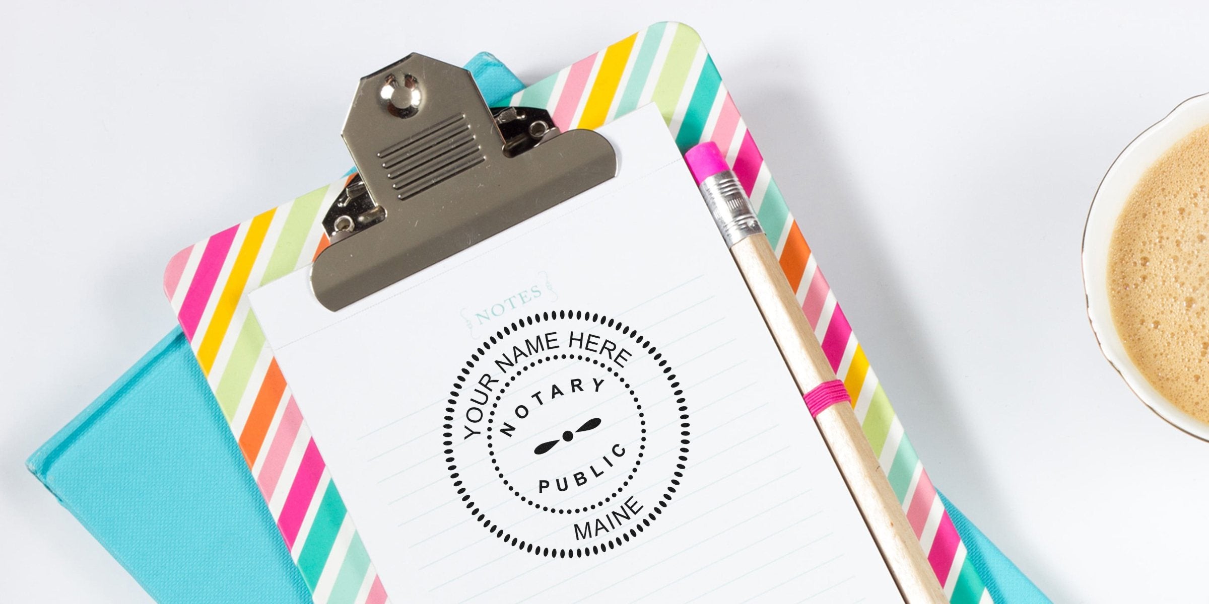 Top-down view of colorful striped clipboard with lined notepad showing "Notary Public" circular stamp, wooden pencil with pink eraser and a cup of frothy coffee on white desk