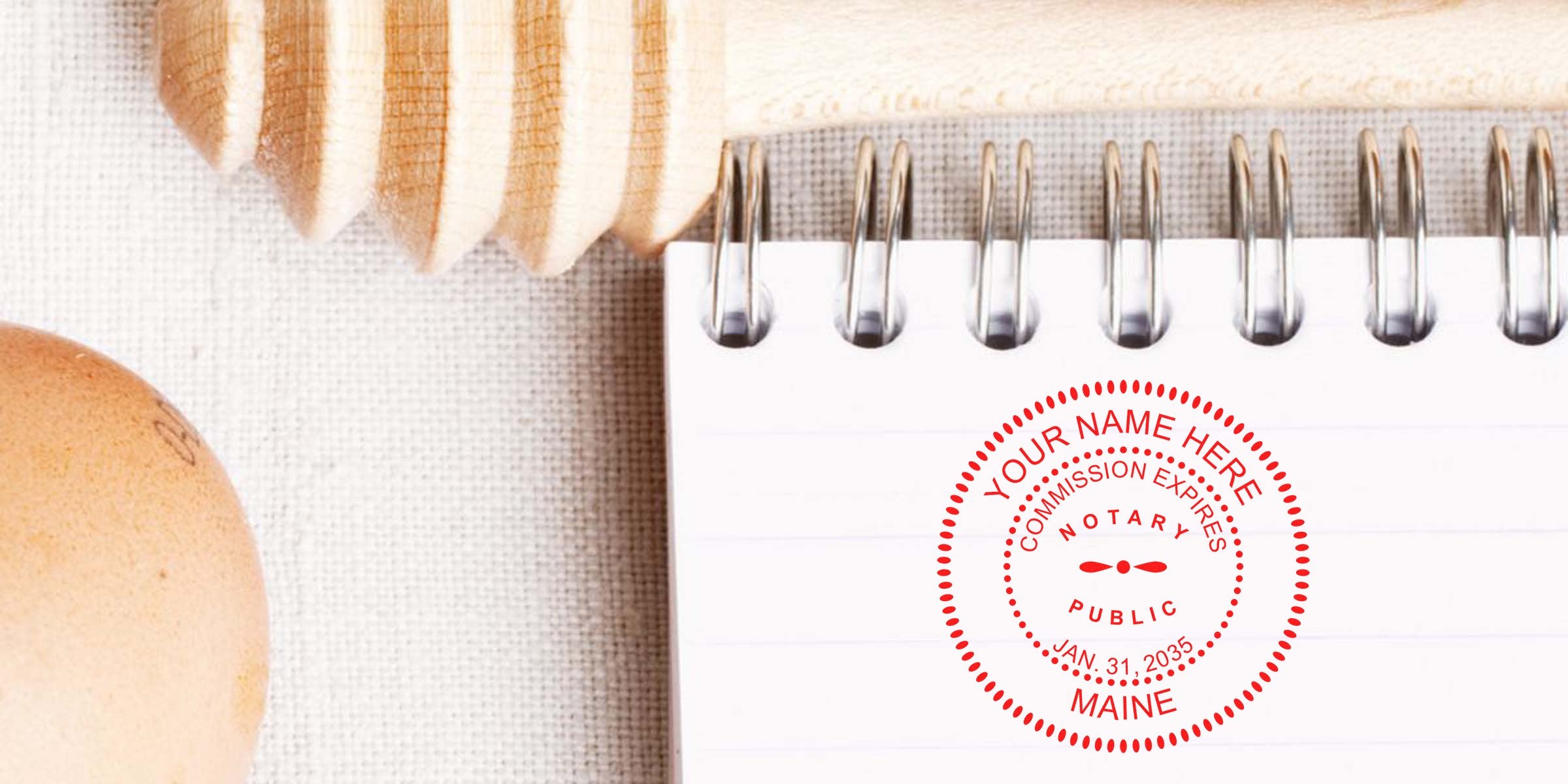 Close-up of a spiral-bound lined notebook page stamped with a red notary public seal reading "Maine," placed on textured fabric beside a wooden honey dipper and a brown egg.