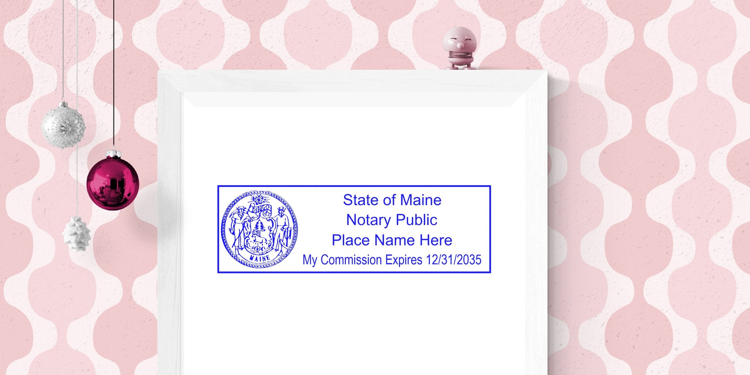 Framed blue State of Maine notary public stamp mockup reading "Place Name Here My Commission Expires 12/31/2035" on pink retro wallpaper with hanging holiday ornaments