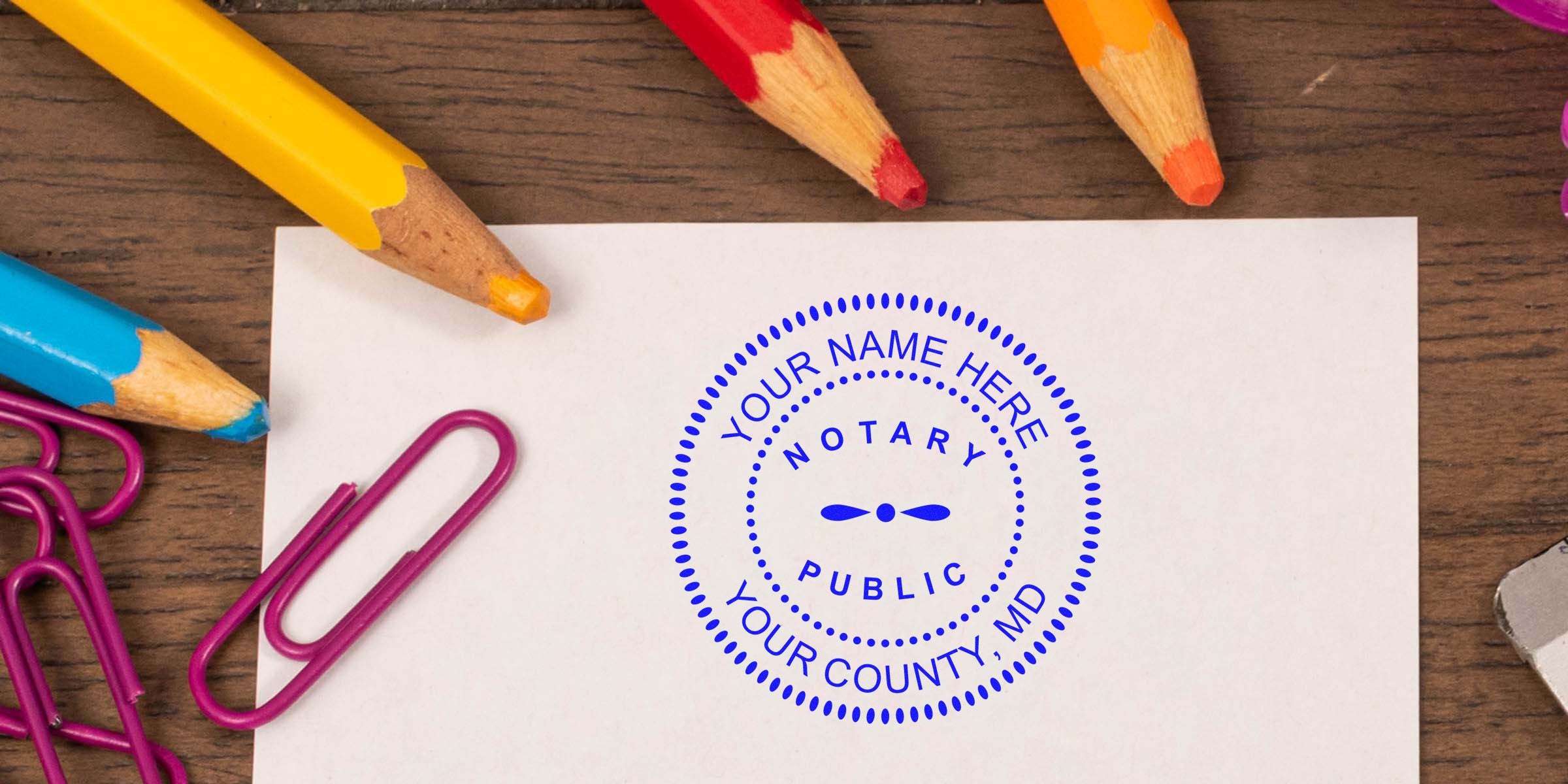 Top-down photo of a blue "Notary Public" custom seal stamped on white paper on a wooden desk, surrounded by colorful pencils and pink paperclips.