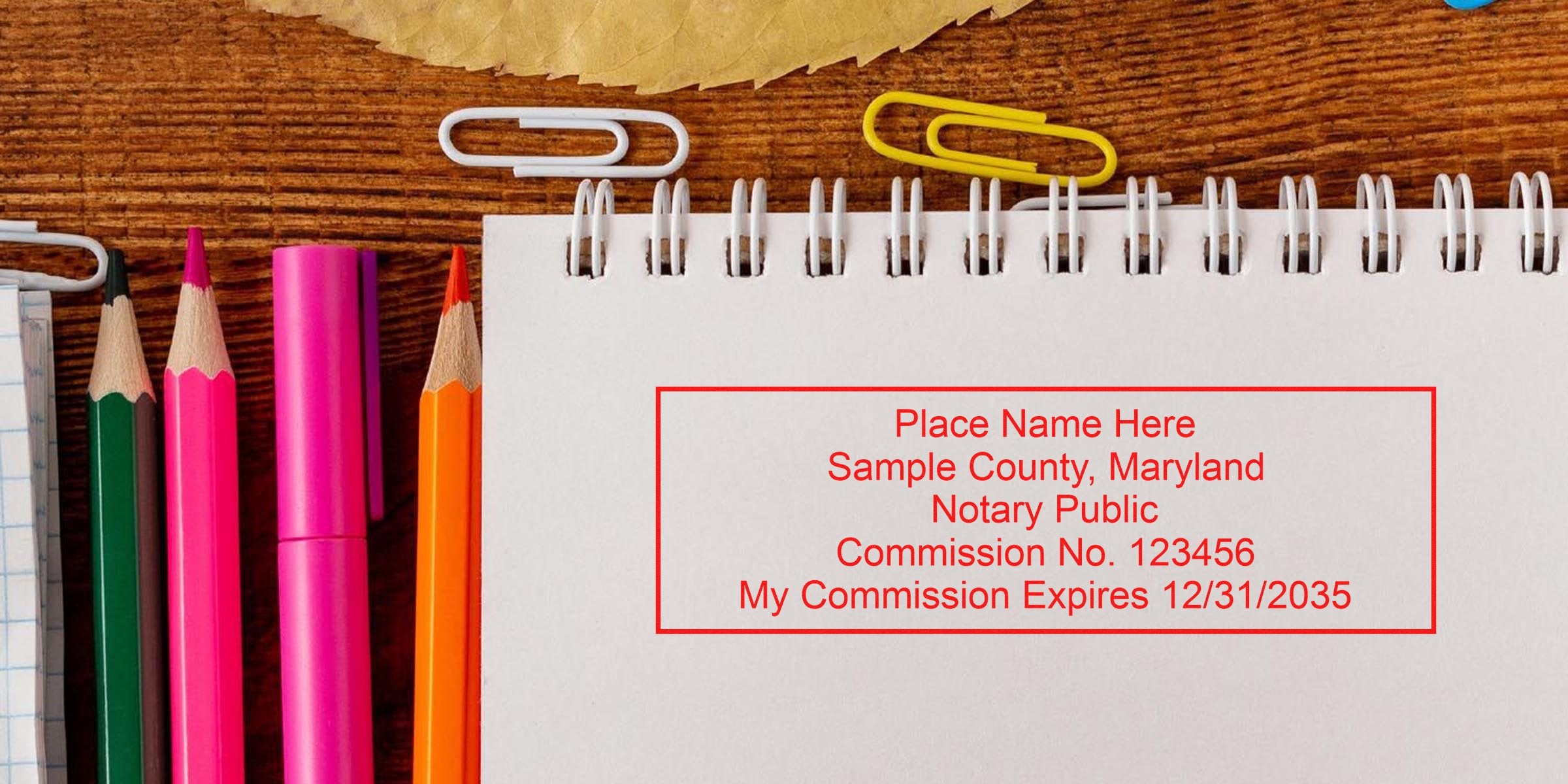 Spiral notebook showing red notary stamp sample text beside colorful pencils, pen and paperclips on a wooden desk — office stationery image.