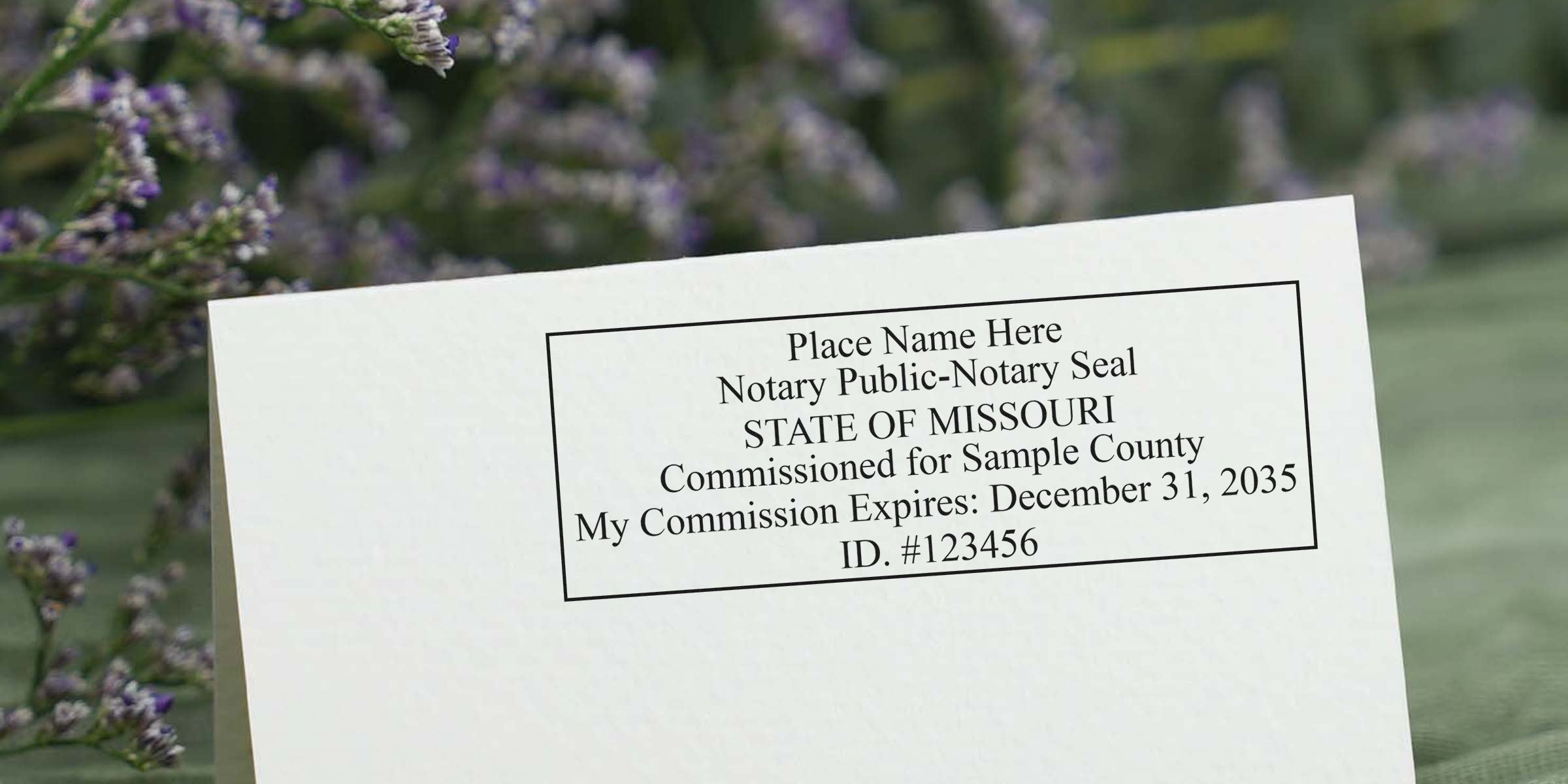 Folded white card displaying a printed notary public seal sample reading "STATE OF MISSOURI" with commission expiration and ID number, soft lavender flowers blurred in background