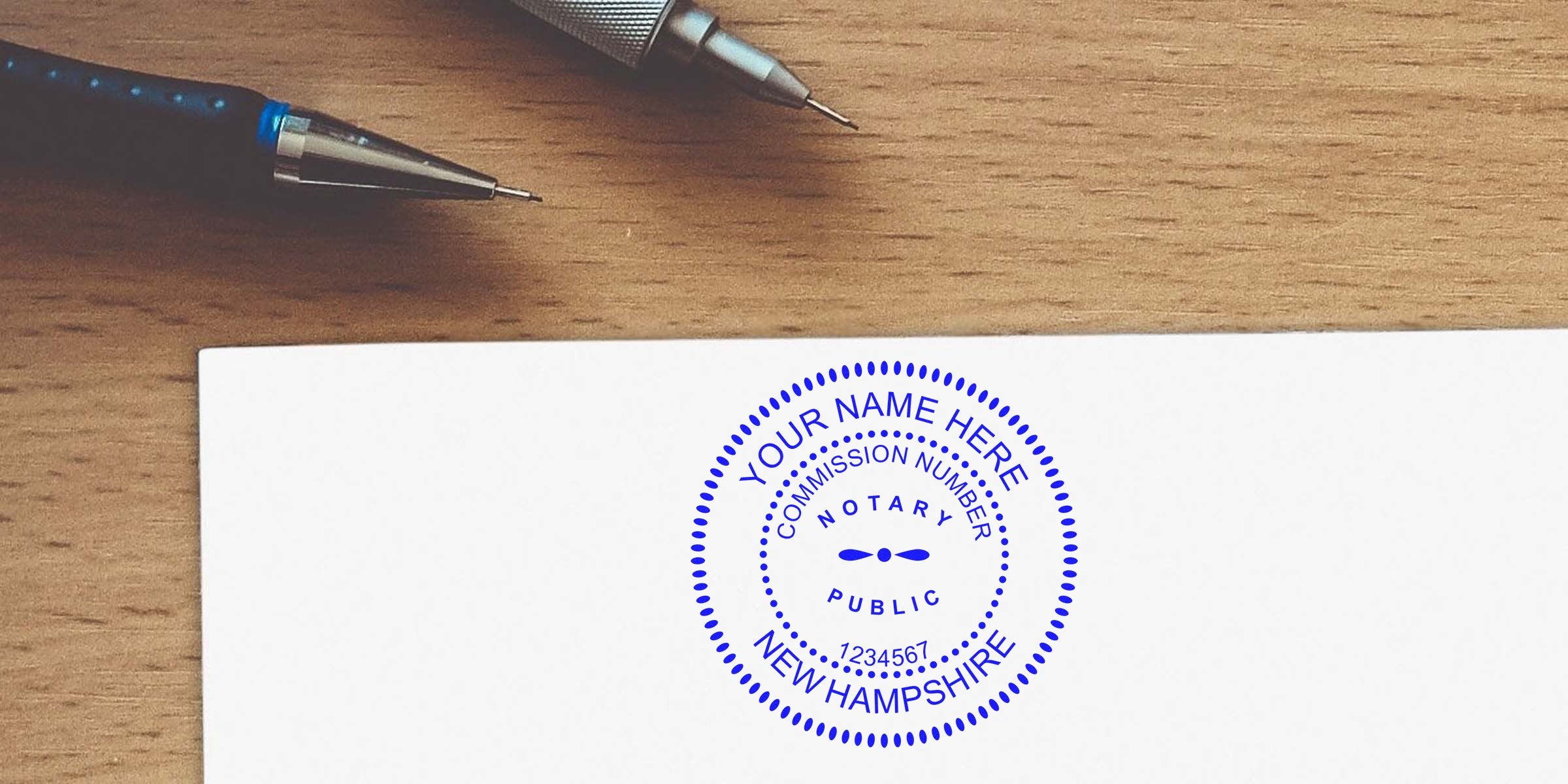 Blue notary public seal stamped on white document reading "New Hampshire" and commission number, with two mechanical pencils on a wooden desk.