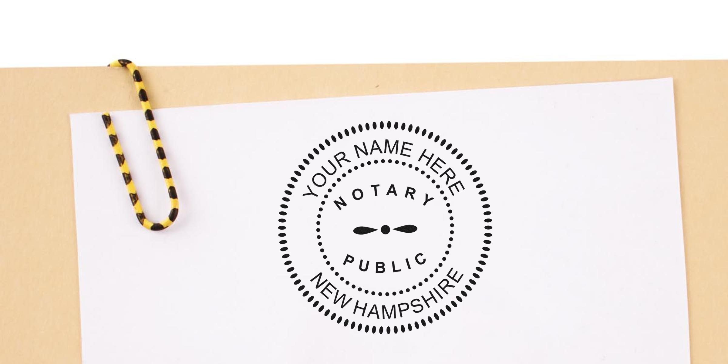 Customizable circular notary public seal stamp reading "Notary Public" and "New Hampshire" on white paper, clipped with a yellow-and-black striped paperclip on a tan folder — notary stamp mockup for branding.