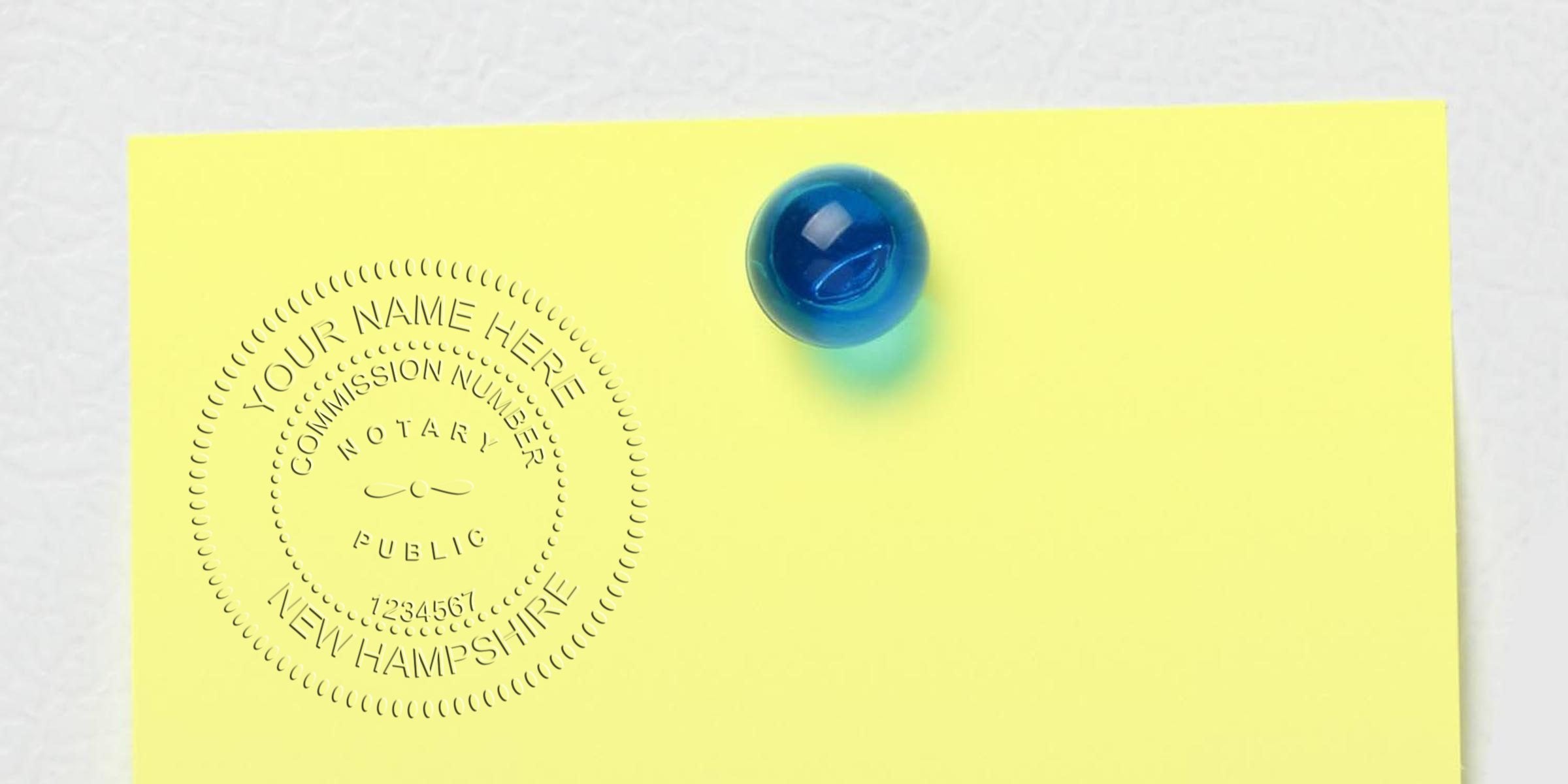 Embossed notary seal on yellow paper reading "Your Name Here, New Hampshire" pinned by a glossy blue glass marble, office stationery close-up