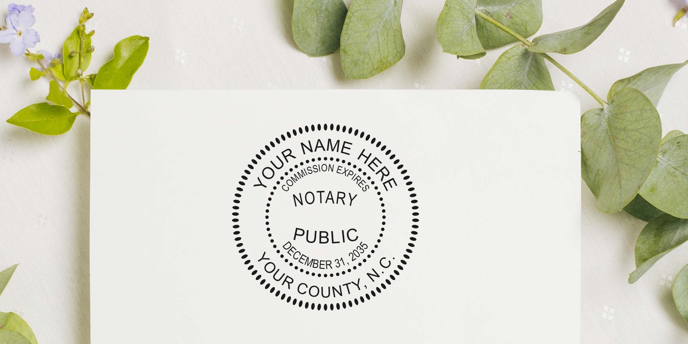 Notary public round stamp mockup on white paper showing customizable seal text and expiration date, flat-lay with green eucalyptus leaves and soft floral background