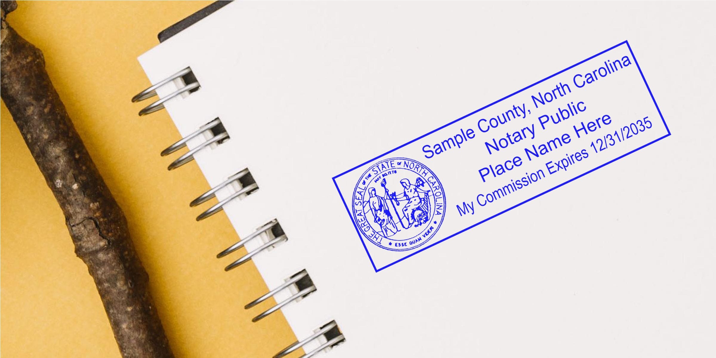 Blue North Carolina notary public stamp on a white spiral-bound notebook page showing sample county, placeholder name and commission expiration date, photographed on a yellow background with a brown twig alongside the notebook.