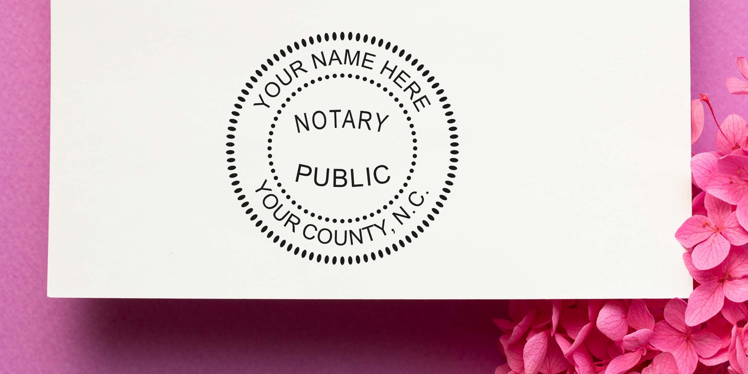 Notary Public circular stamp mockup on white card with placeholder text "Your Name Here, Your County, N.C." on pink paper background and pink hydrangea flowers — notary service business stationery concept.