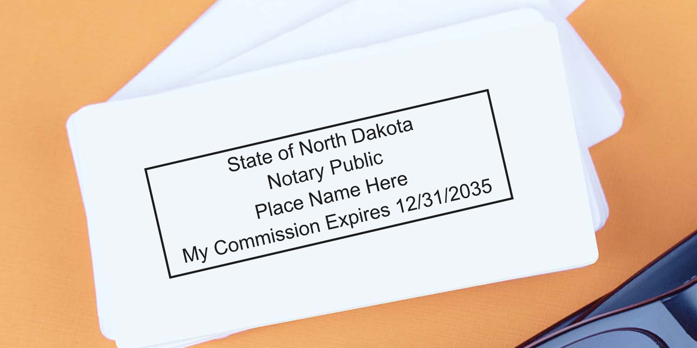 Stack of white notary commission cards on an orange desk showing a rectangular stamp reading "State of North Dakota Notary Public Place Name Here My Commission Expires 12/31/2035" with eyeglasses at the corner