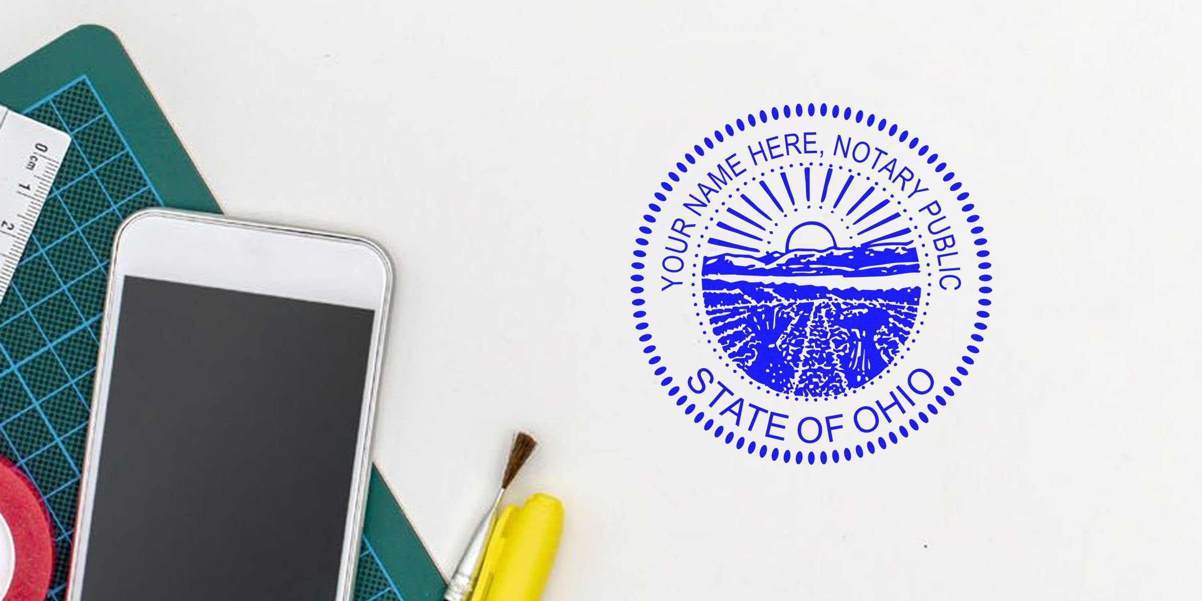 Blue State of Ohio notary stamp on white desk beside smartphone, cutting mat, ruler, paintbrush and yellow highlighter — office workspace image