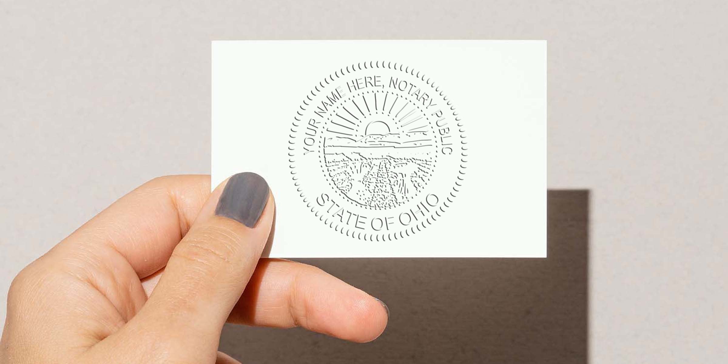 Hand holding small white card with embossed circular State of Ohio notary public seal depicting sunrise and landscape, neutral background, close-up of fingers with gray nail polish.