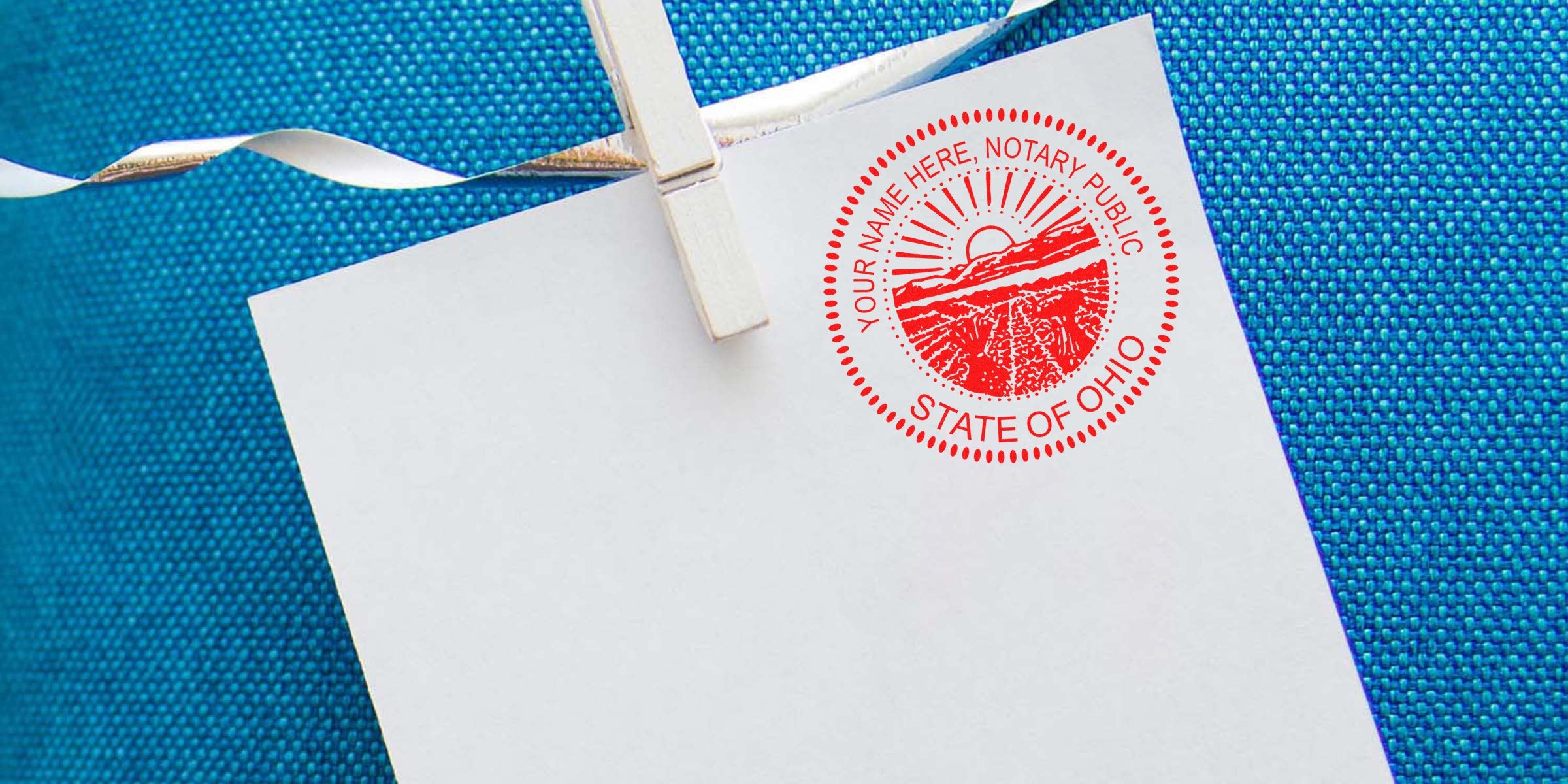 Blank white paper clipped with a wooden clothespin showing a red "State of Ohio" notary seal stamp on a textured blue fabric background, close-up of notary stamp for documents and legal certification.