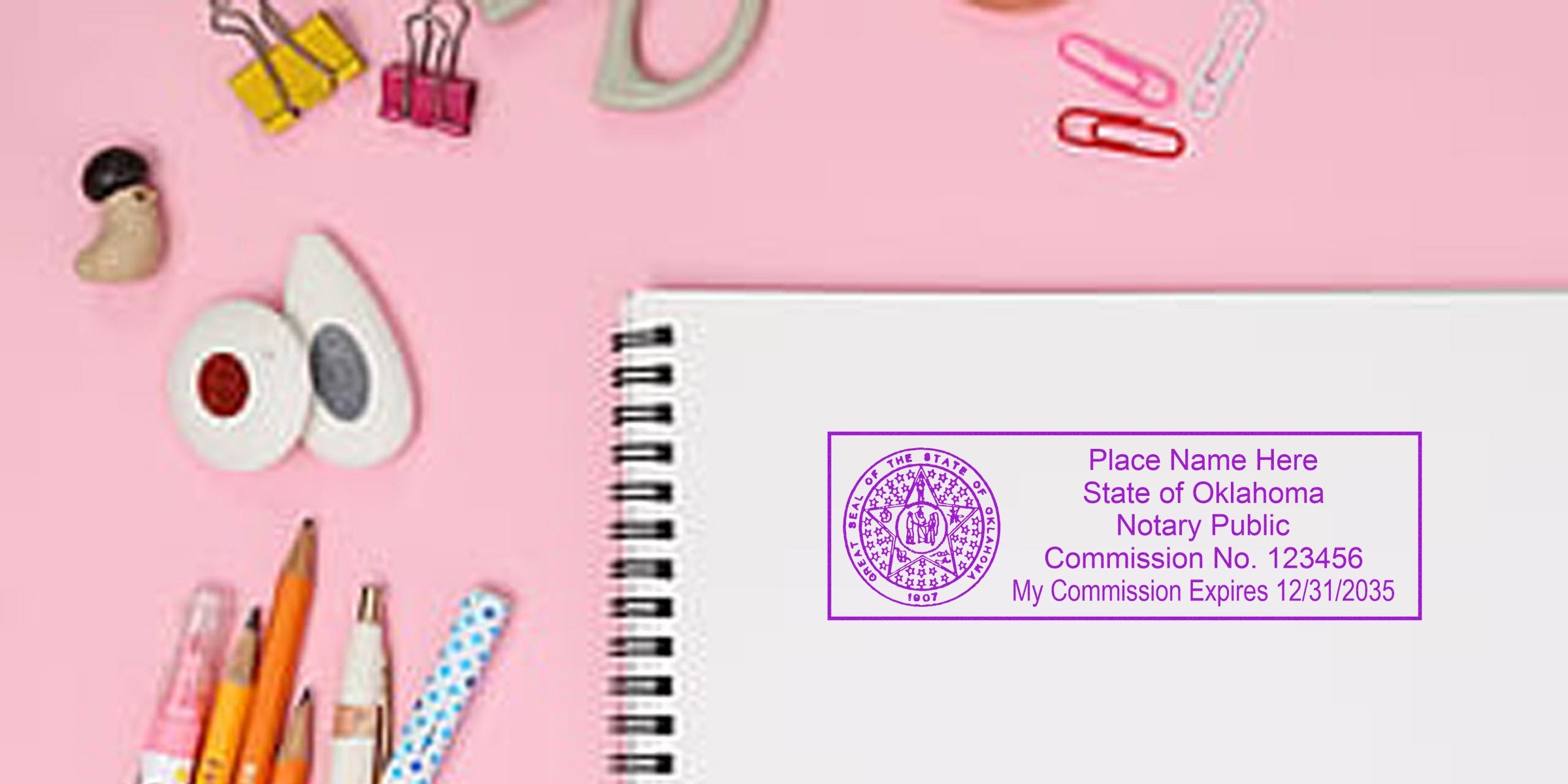 Flat lay pink desk with spiral notebook showing a purple Oklahoma notary public stamp, pencils, pens, decorative tape, paper clips and binder clips