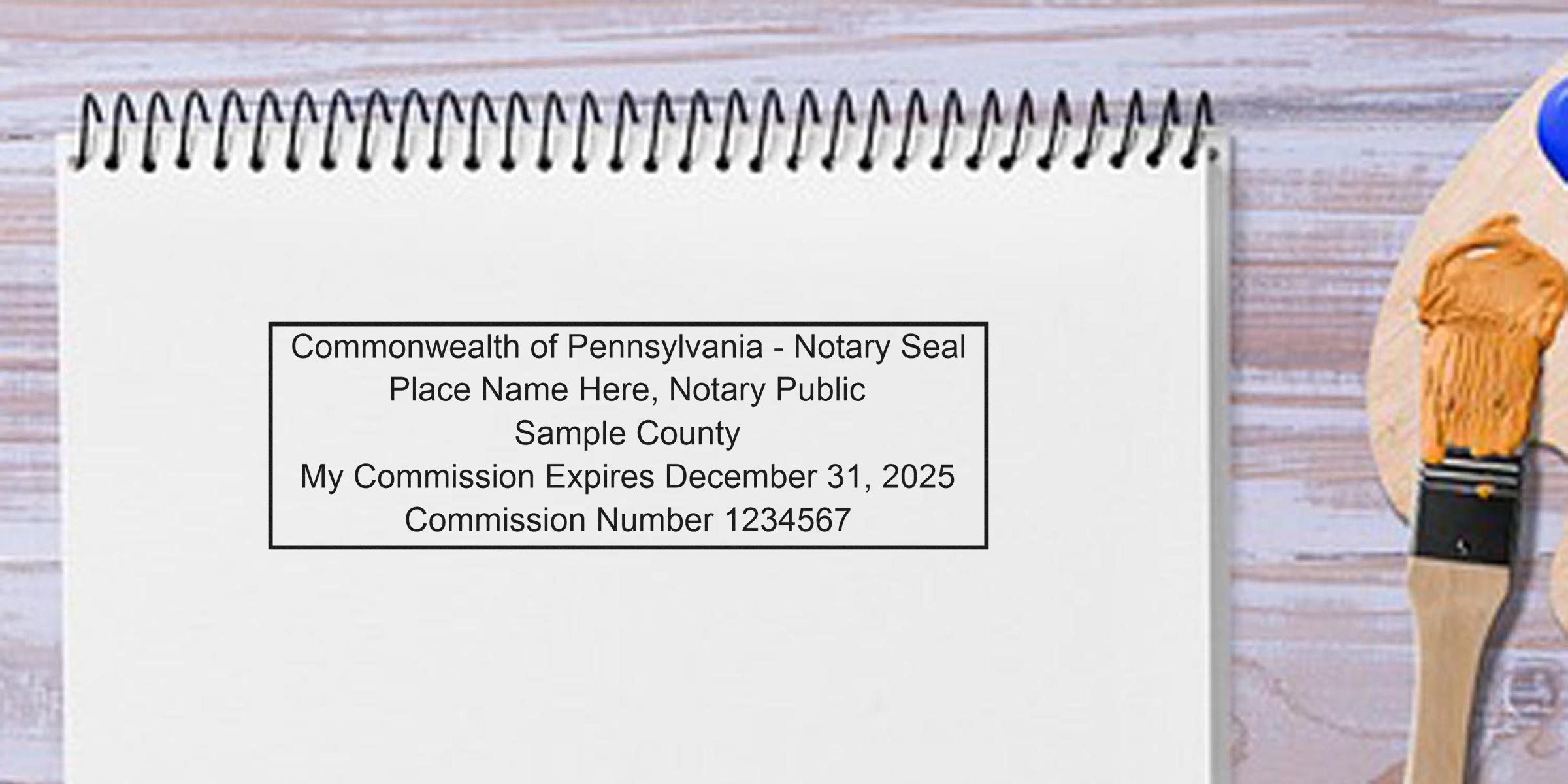 Mockup of a Commonwealth of Pennsylvania notary seal stamp on a spiral notebook page reading sample county, commission expires December 31, 2025, commission number 1234567, paintbrush nearby