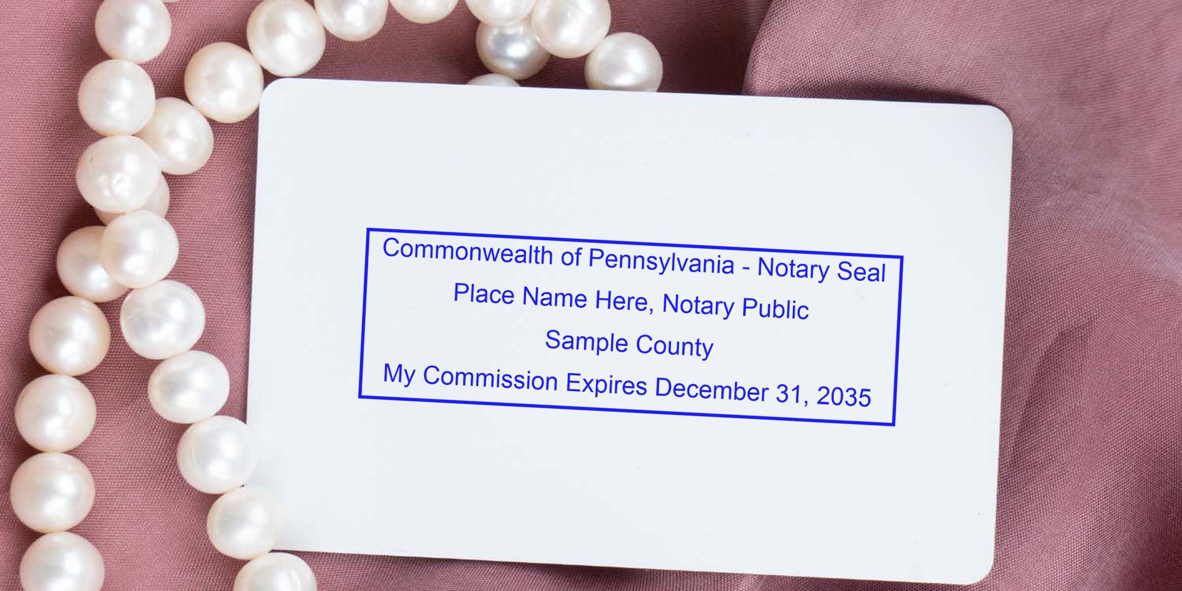 White card showing blue Pennsylvania notary seal sample text with commission expiration date, set on pink fabric beside a string of pearls