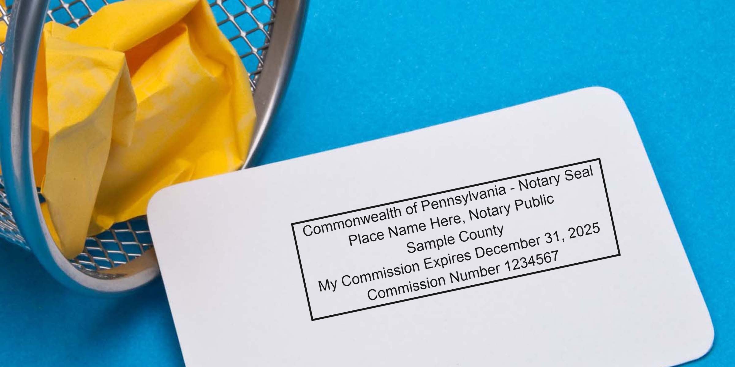 White card showing a sample Pennsylvania notary seal and commission details, placed on a bright blue background beside a small metal wastebasket containing crumpled yellow paper.