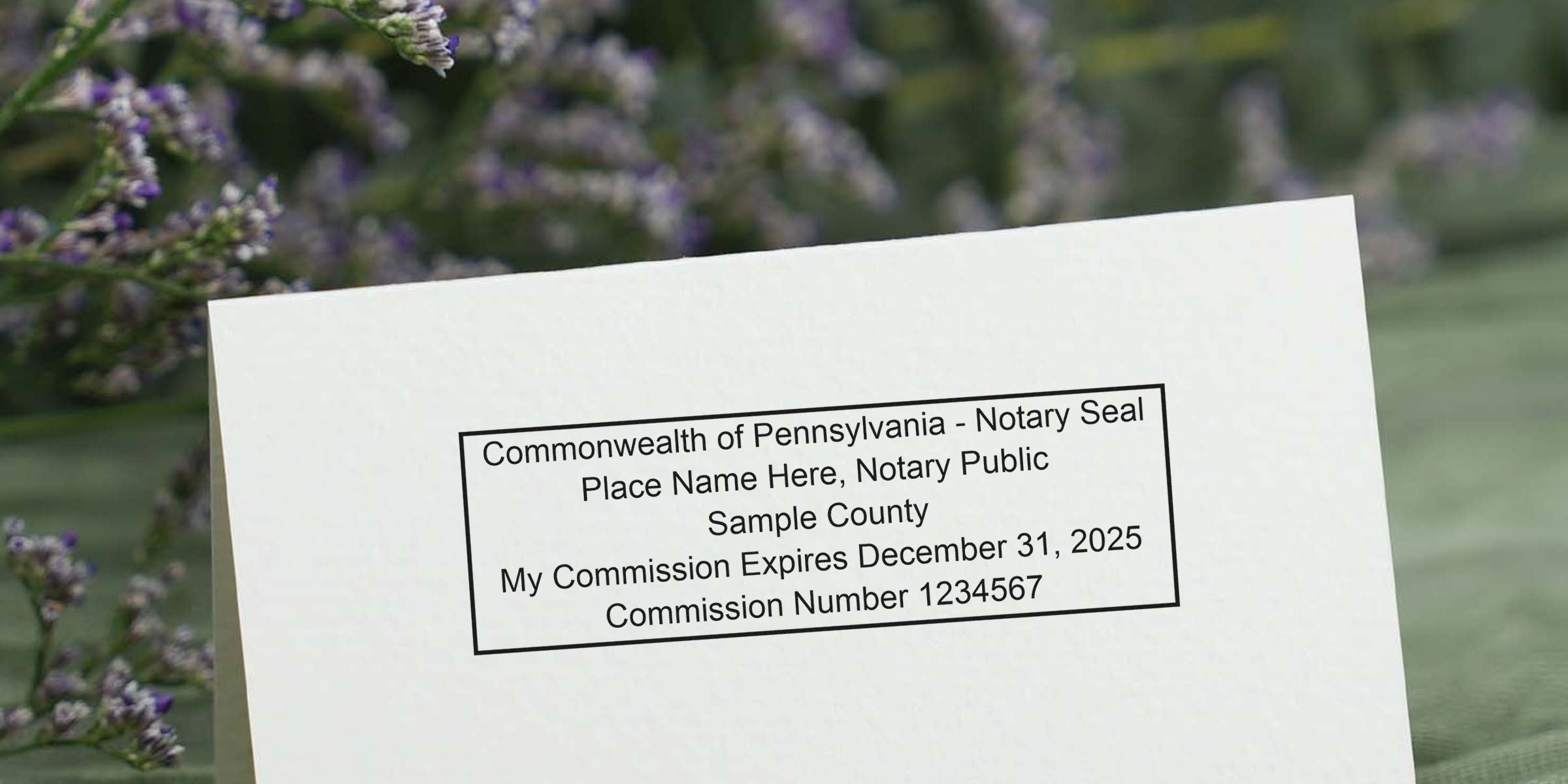 Sample Commonwealth of Pennsylvania notary seal printed on white card with placeholder text and commission details, lavender flowers blurred background