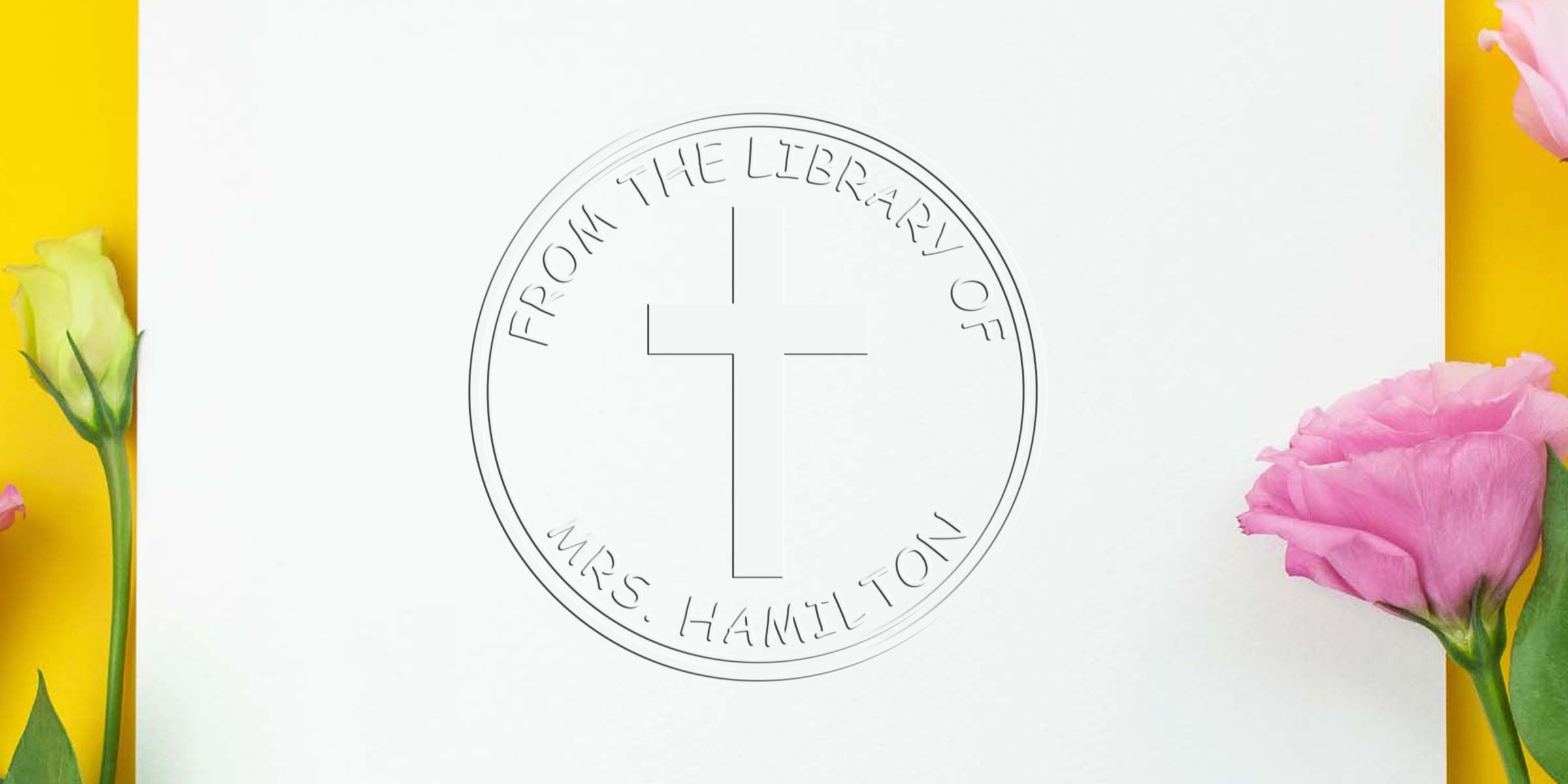 Round coin/medallion with a cross in the center and the inscription “From the Library of Mrs. Hamilton” around the edge, placed on a white background with yellow borders and pink flowers on the sides.