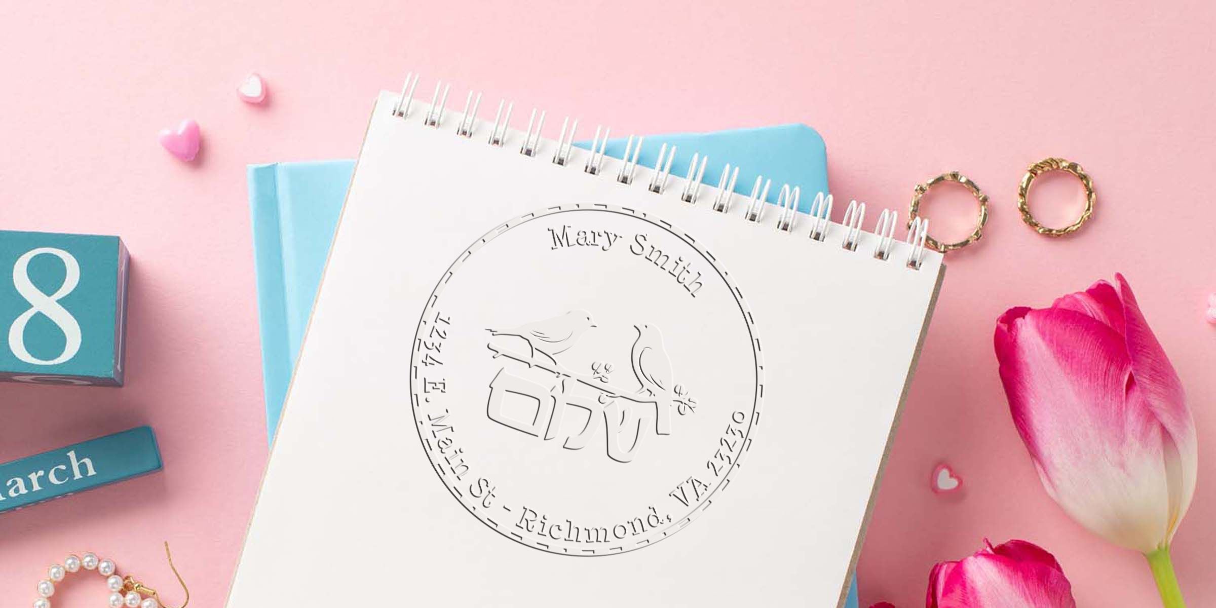 Notebook page with a circular stamp design of two birds on a bench, surrounded by text “Mary Szech” and “Richmond, VA,” on a pink desk with blue blocks and pink tulips.