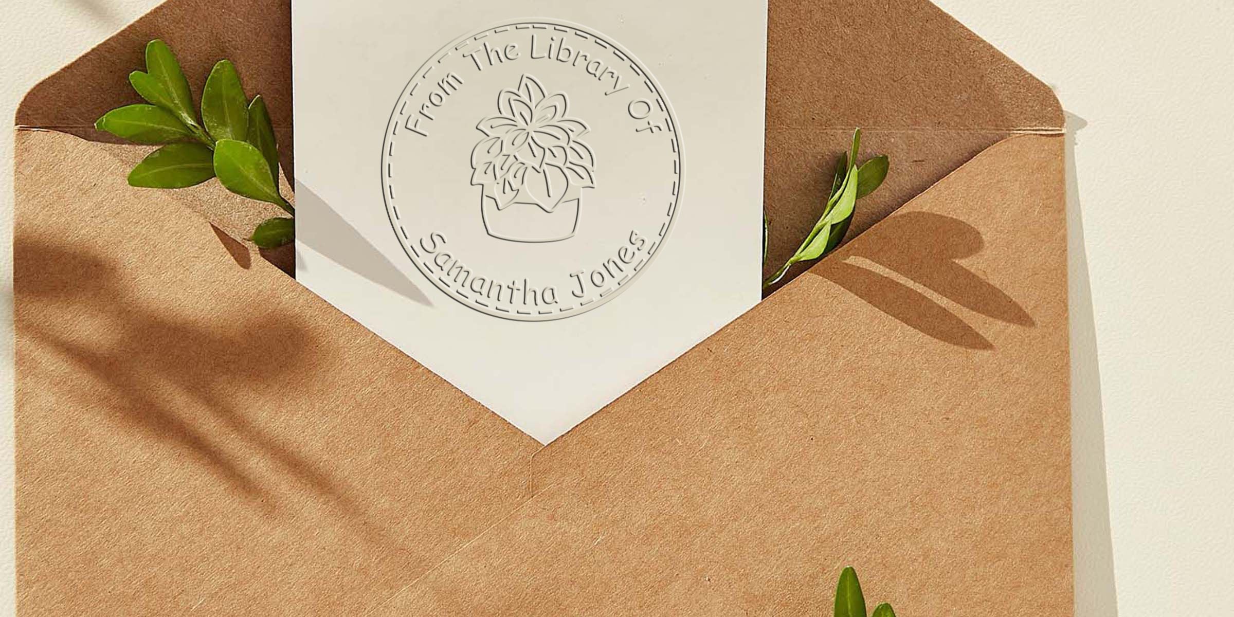 A white card with a circular embossed seal reading “From The Library Of Samantha Jones” sits in a brown envelope with green leaves peeking out.