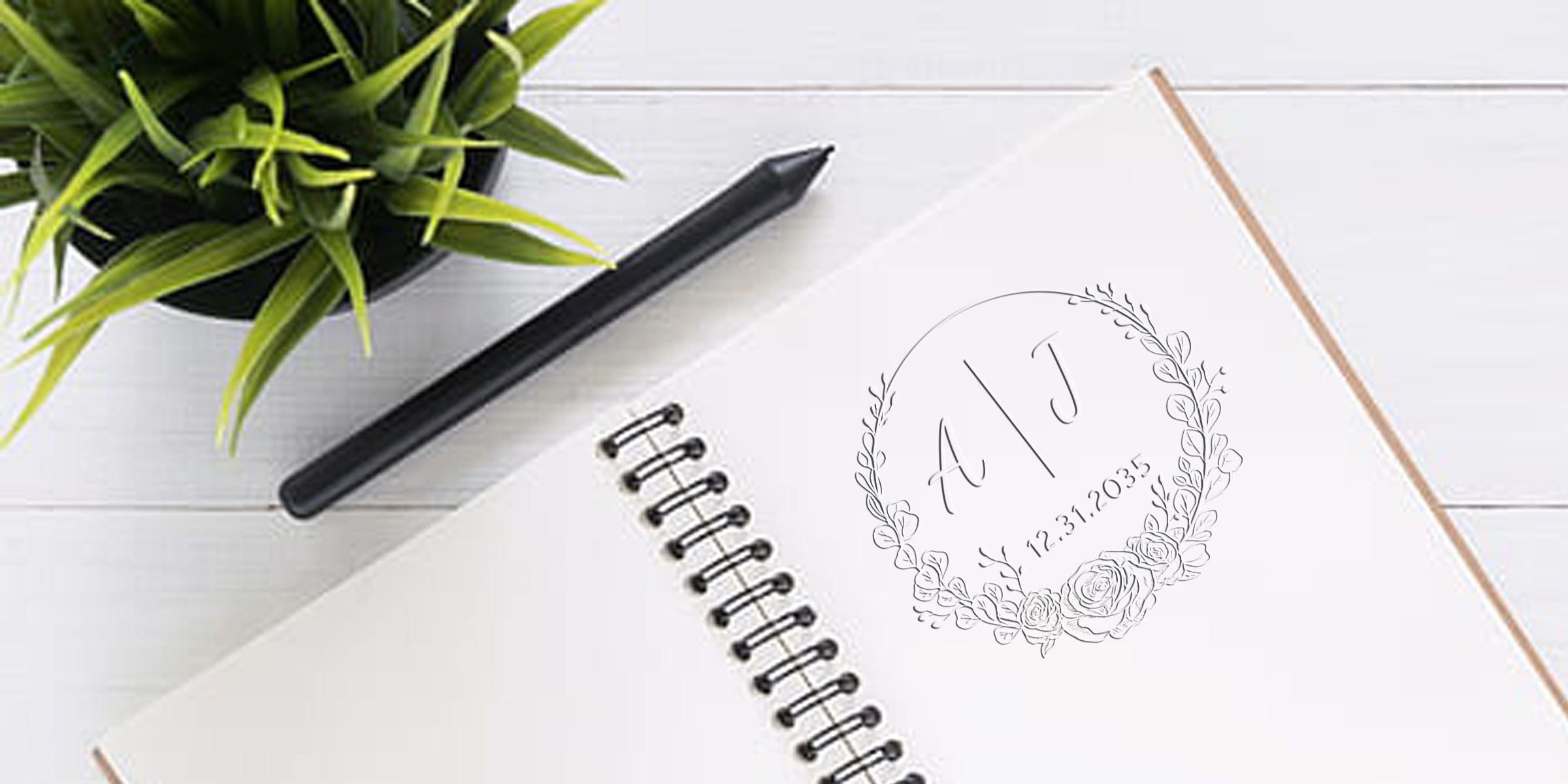 Notebook page with a floral wreath monogram “A J” and date 12.31.2035, beside a pencil and a potted plant on a white wooden desk.
