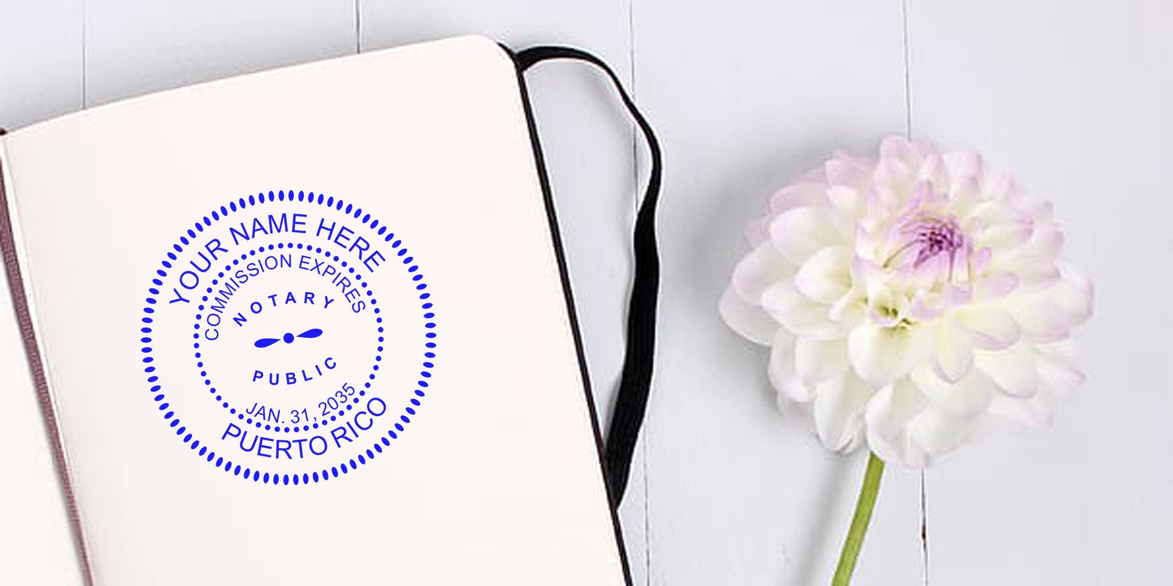 Open notebook showing blue circular Puerto Rico notary stamp mockup on blank page beside pale pink-white dahlia flower on white wooden surface