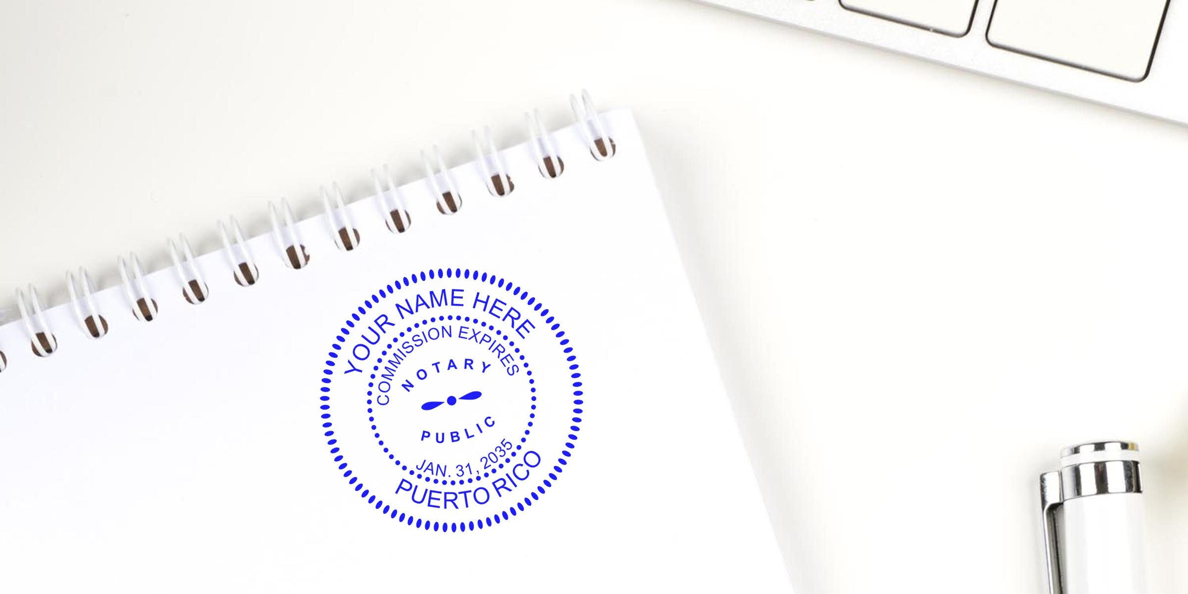 Blue circular notary public seal stamp reading "Puerto Rico" on a white spiral notebook next to a silver pen and laptop keyboard on a clean desk