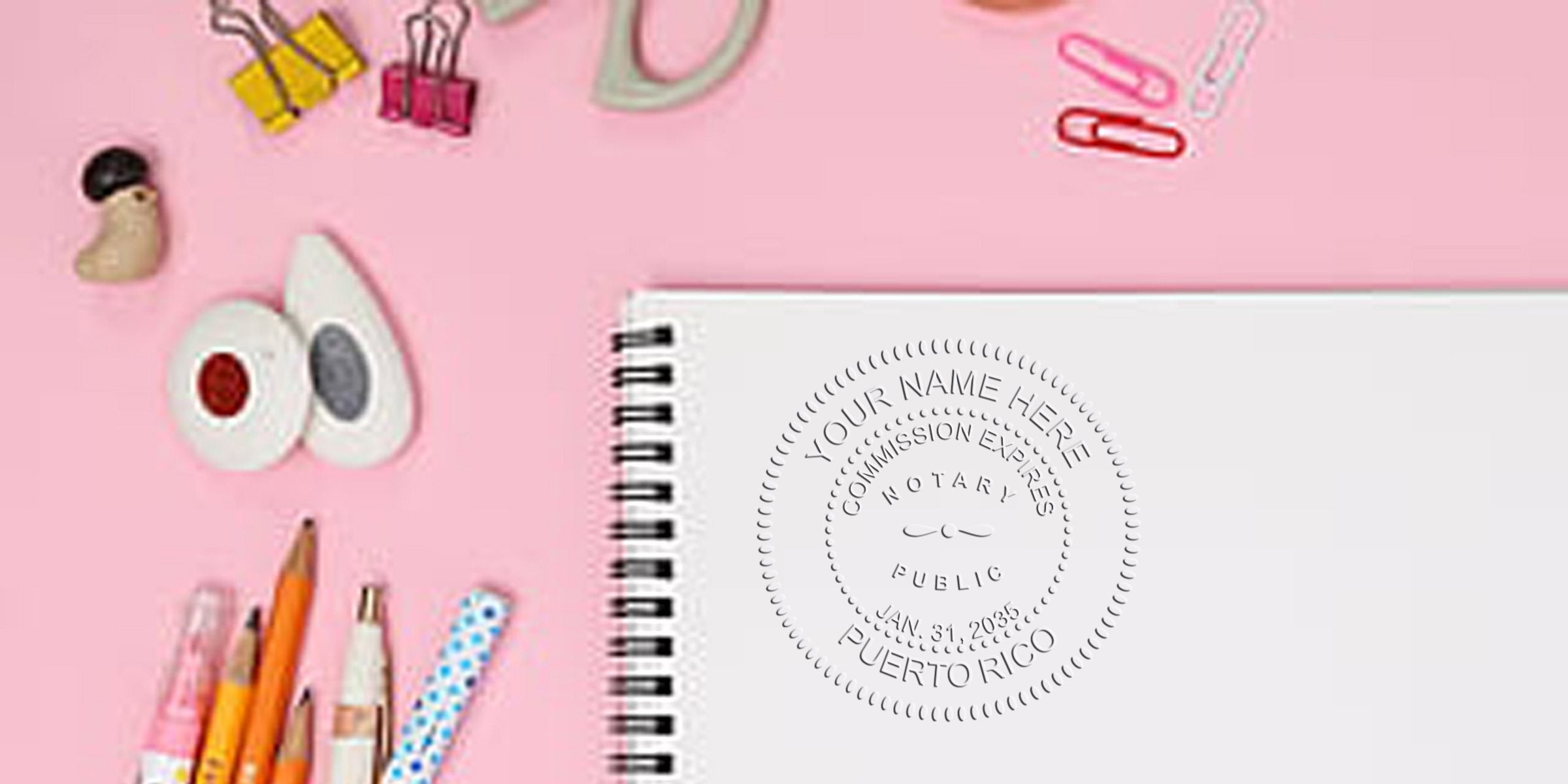 Flat-lay pink desk with pencils, pens, binder clips, decorative tape and a spiral notebook showing an embossed circular notary public seal reading Your Name Here, commission expires, Puerto Rico.