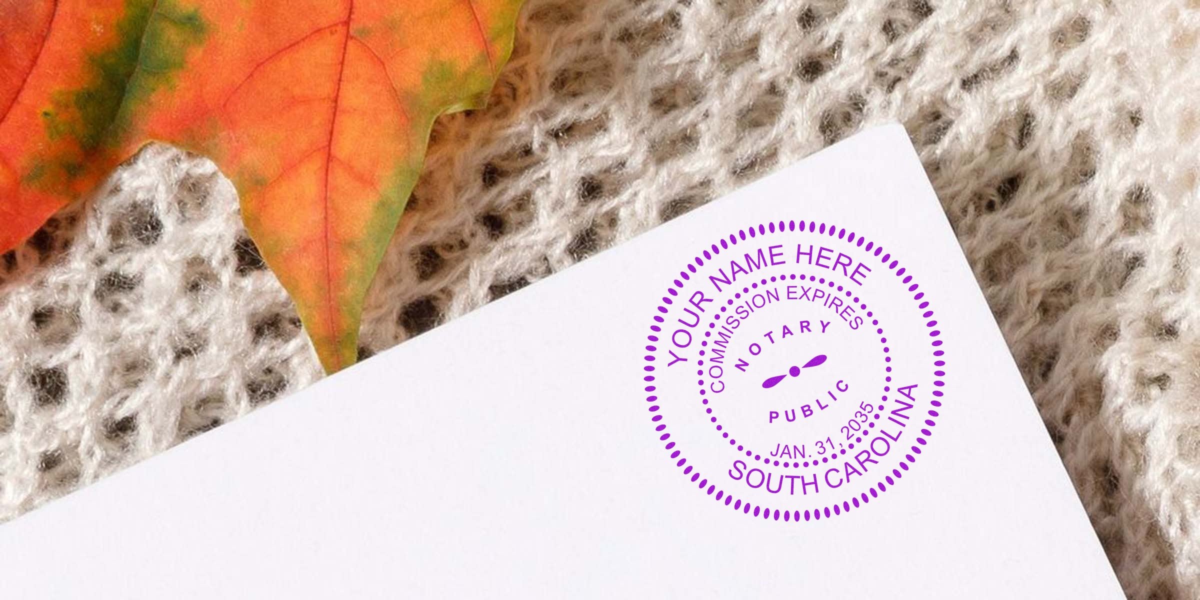 Close-up of an orange autumn leaf on a cream knitted sweater beside a white envelope stamped with a purple circular South Carolina notary seal.