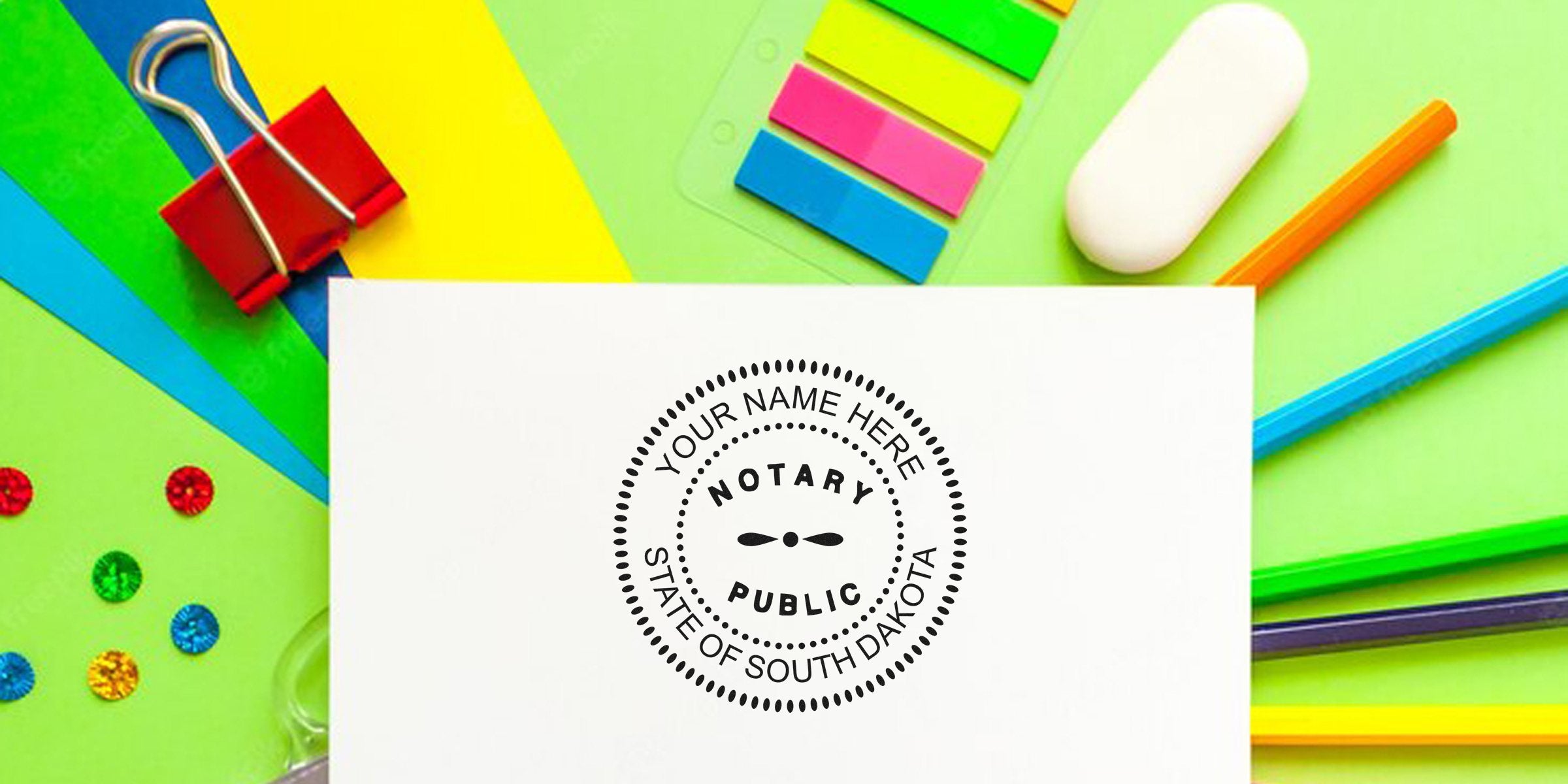 Notary public round stamp mockup reading "Your Name Here, State of South Dakota" on white paper, surrounded by colorful stationery (pencils, sticky flags, binder clip, eraser) on bright background