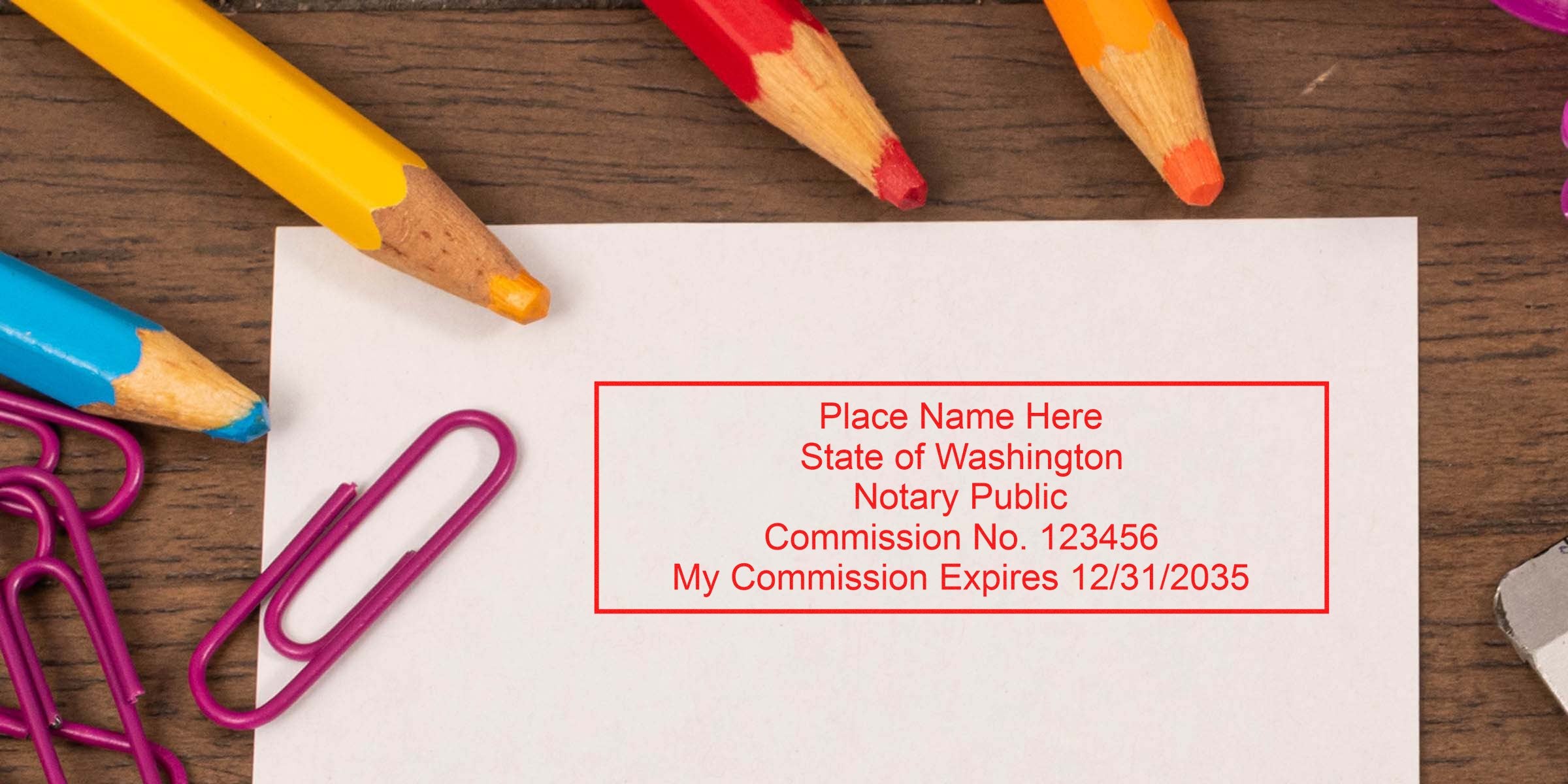 Top-down photo of white paper on a wooden desk with colorful pencils and purple paperclips, featuring a red Washington notary public stamp sample with commission number and expiration date.