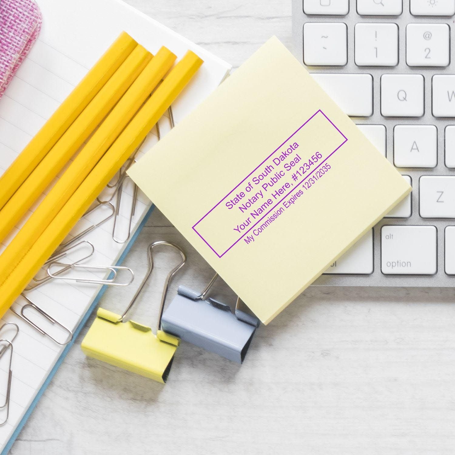 Alt Text: South Dakota Notary Supplies Blog Post Feature Image: Notary stamp on a sticky note beside a keyboard, paper clips, and pencils.