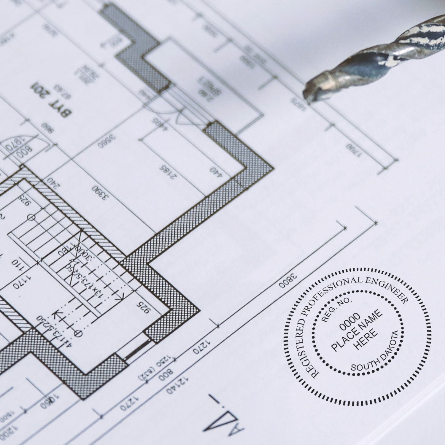 Blueprint with a South Dakota Engineer’s Seal Stamp in use, featuring detailed architectural plans and a drill bit in the background.