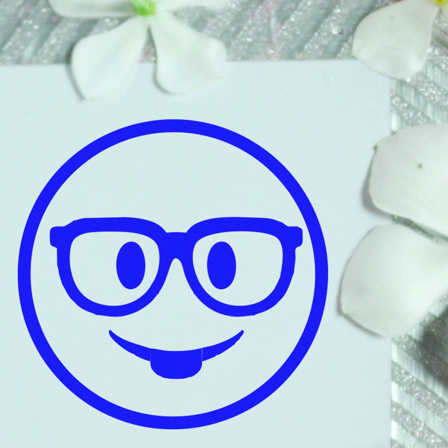 Self-Inking Smarty McGlasses Emotes Stamp featuring a blue smiley face with glasses and a playful tongue, surrounded by white petals on a textured surface. Perfect for adding fun to any project.