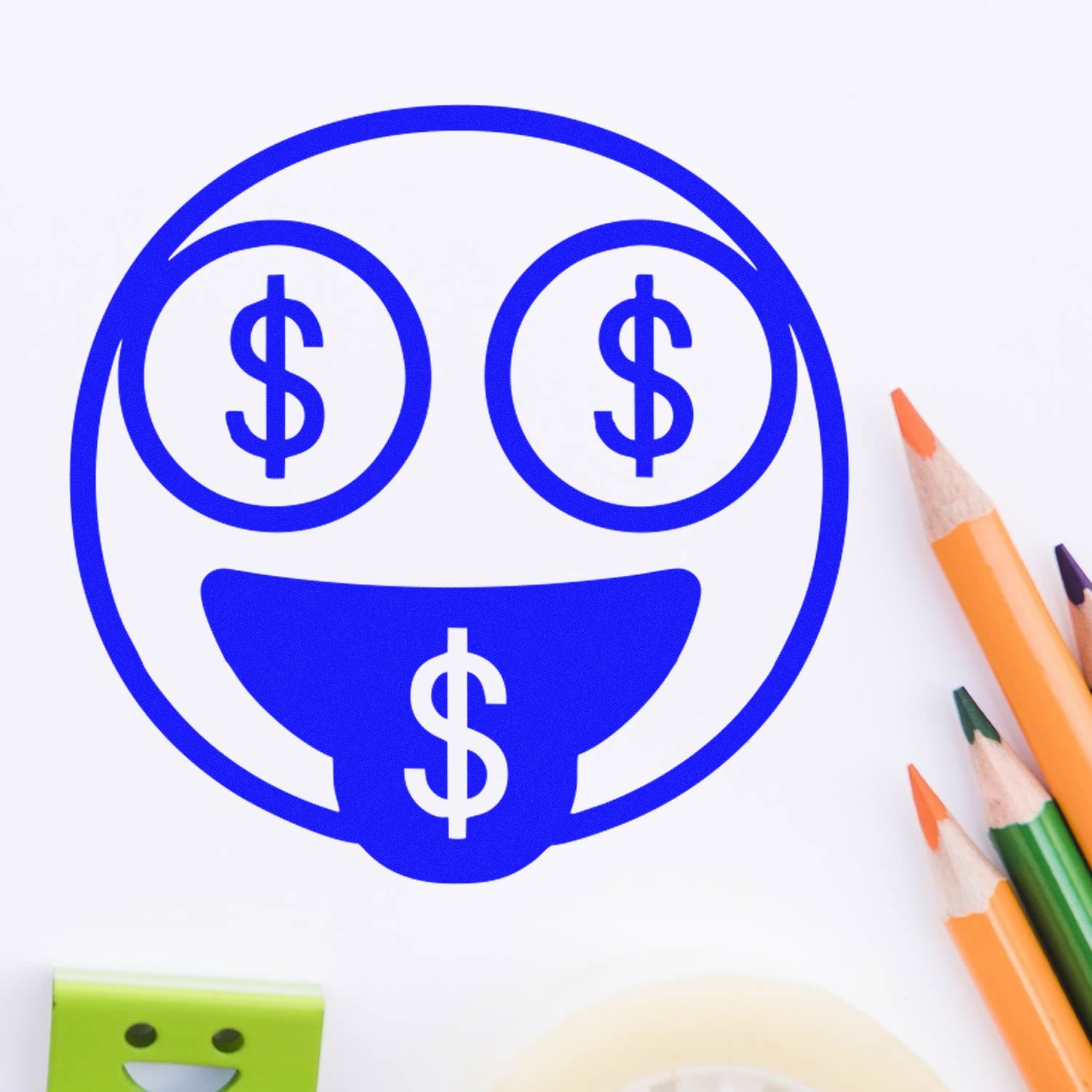 Self-Inking Money Eyes Facial Expressions Stamp in blue, featuring a smiling face with dollar signs for eyes and tongue. Includes colorful pencils and tape in the background.