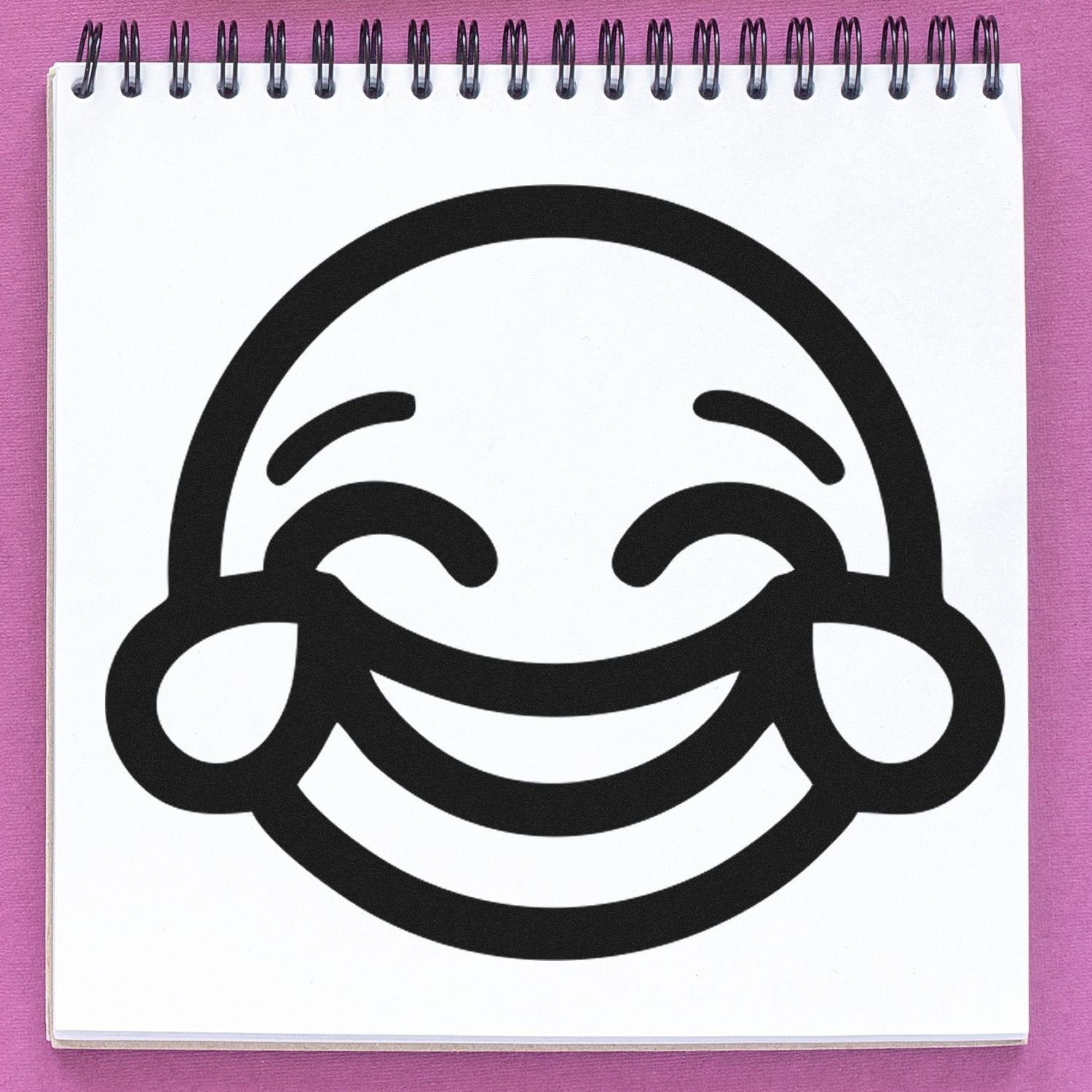 Self-Inking Cheeky Grin Emoji Stamp on a spiral notebook, featuring a bold, smiling face with closed eyes and rosy cheeks. Perfect for adding fun to notes and crafts.