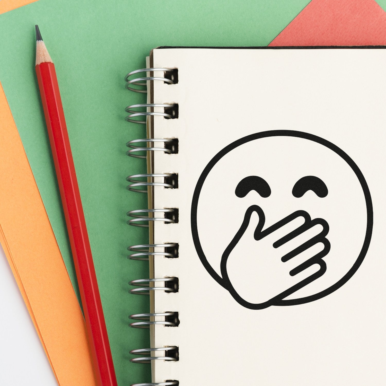 Self-Inking Giggling Gus Emoji Stamp on a notebook, featuring a laughing face with a hand over its mouth. Includes a red pencil and colorful paper background. Perfect for fun, creative stamping.