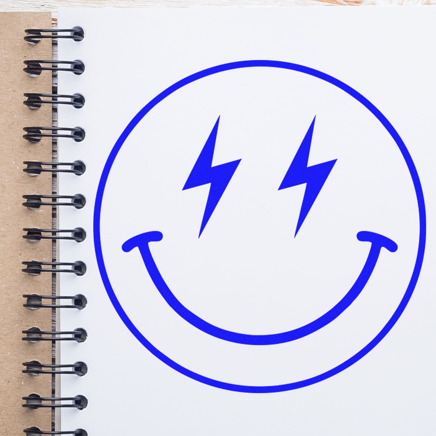 Self-Inking Electric Joy Emoji Stamp imprint on notebook page, featuring a blue smiling face with lightning bolt eyes. Perfect for adding fun and personality to your notes.