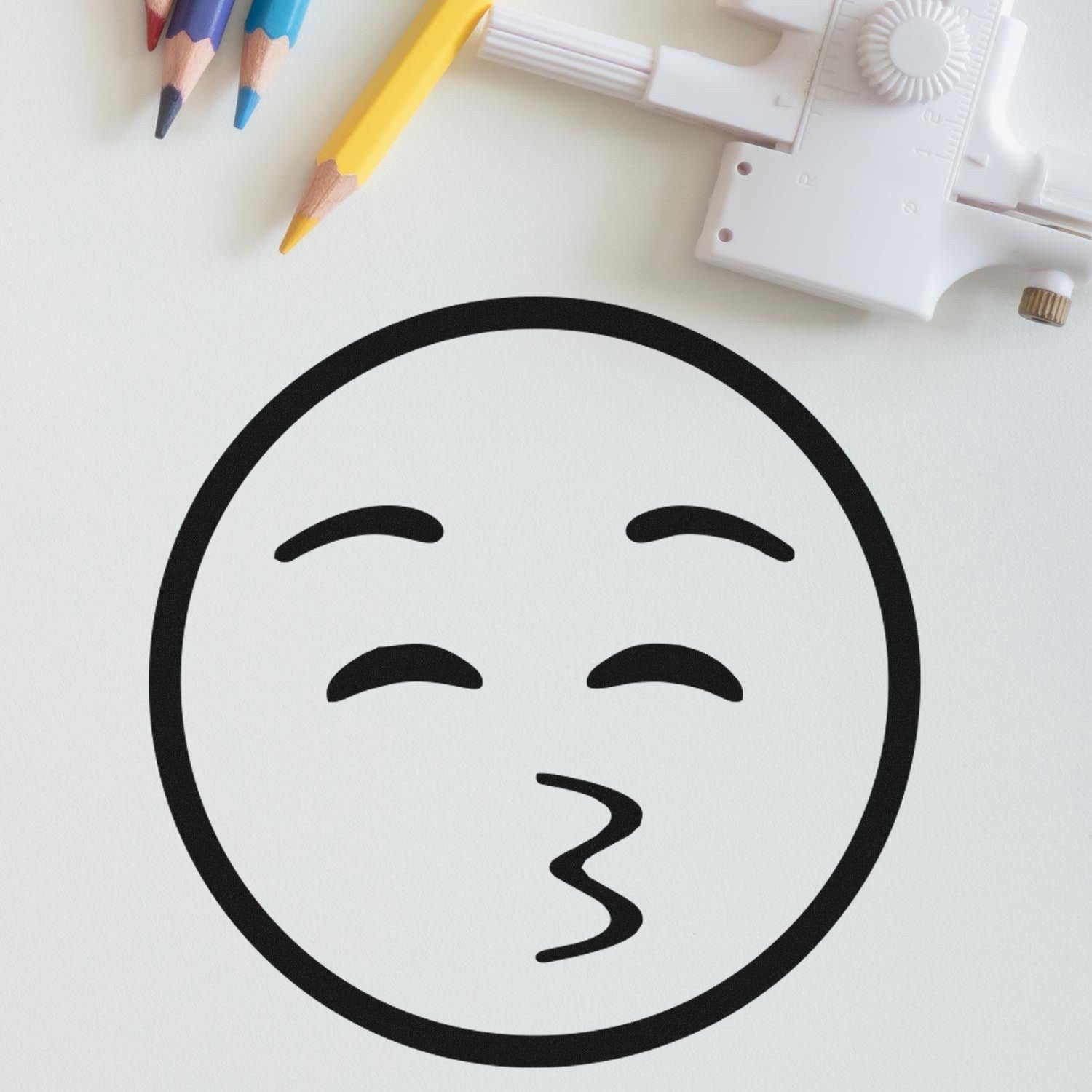 Self-Inking Whistling Willie Emoji Stamp on paper, showcasing a playful whistling face. Ideal for adding fun to notes and crafts. Includes pencils and a white tool in the background.