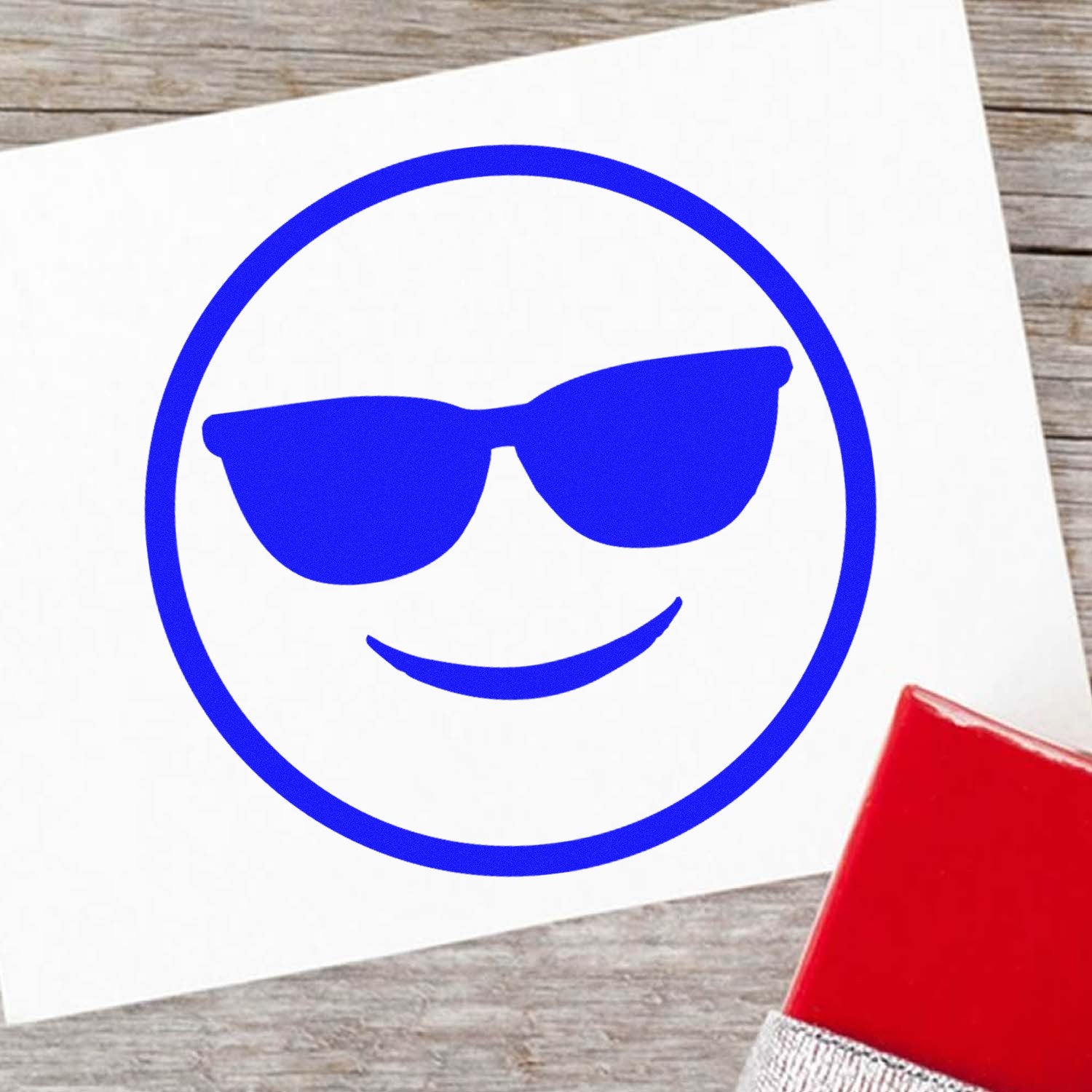 Wood Handle Cool Carl Emoji Rubber Stamp with a blue smiling face wearing sunglasses, displayed on a wooden surface with a red ink pad.