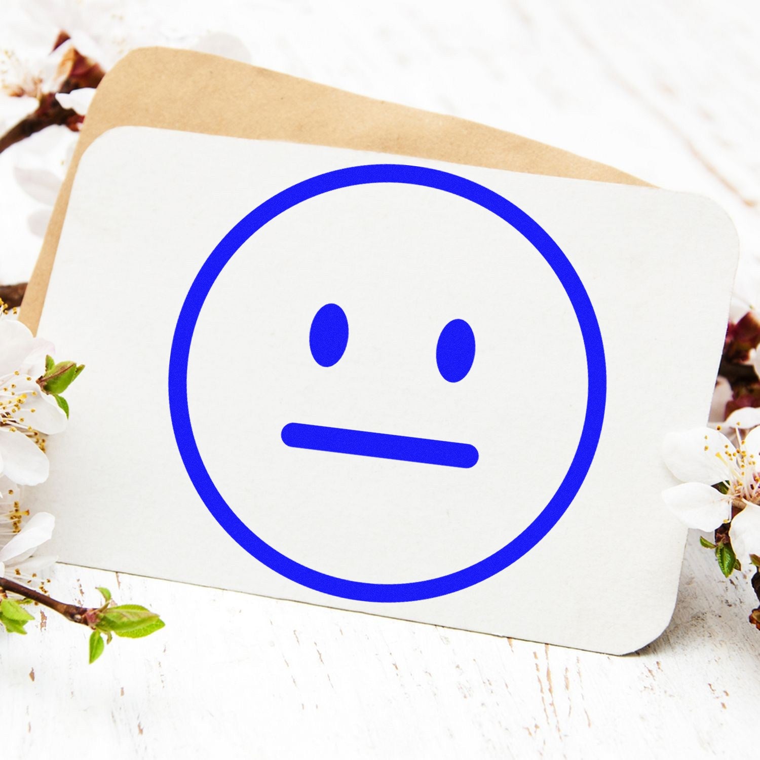 Self-Inking Neutral Ned Emoji Stamp with a blue neutral face design on a white card, surrounded by delicate white flowers on a light wooden surface.