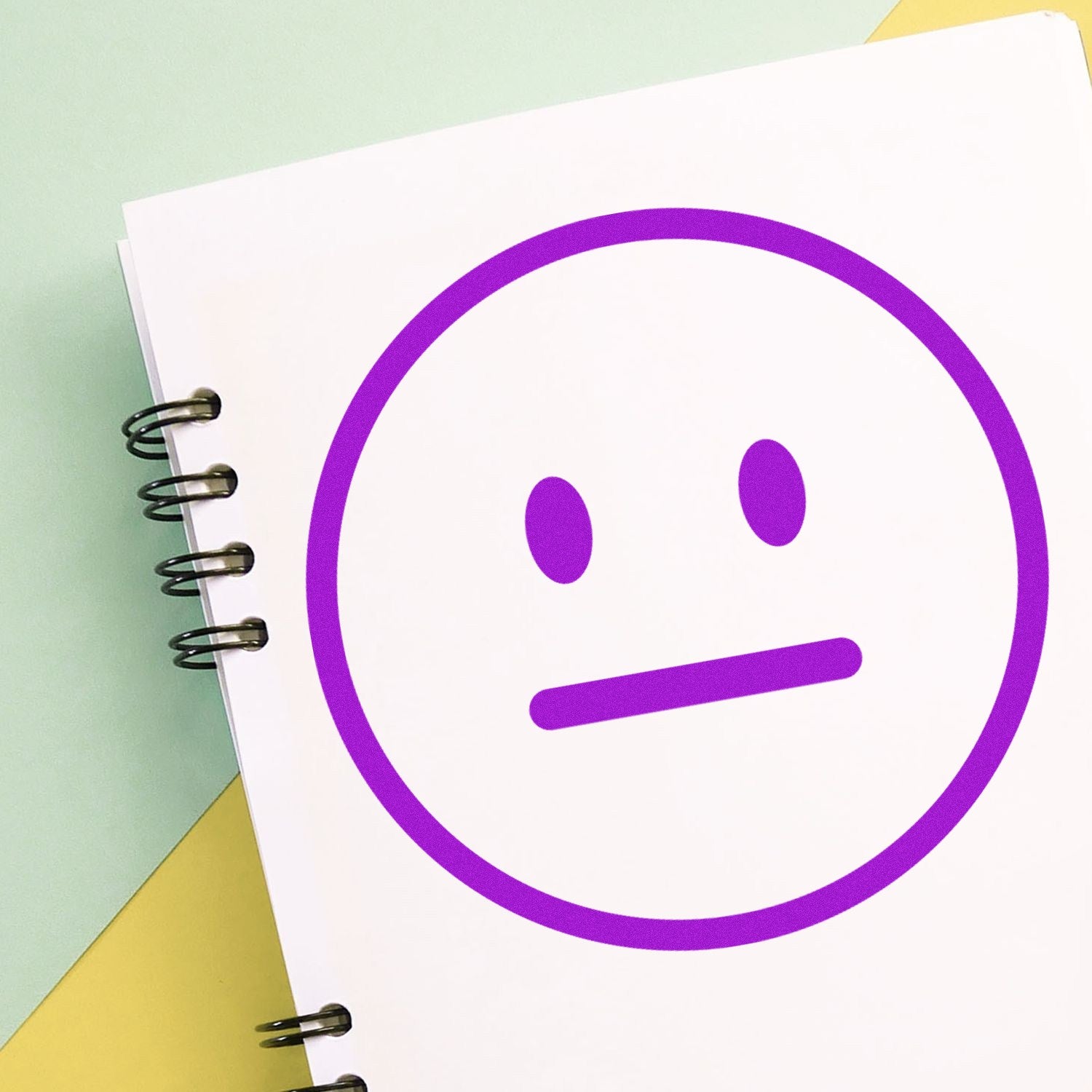 Self-Inking Neutral Ned Emoji Stamp with a purple neutral face design on a spiral notebook, showcasing its crisp and clear imprint. Perfect for adding personality to notes and documents.