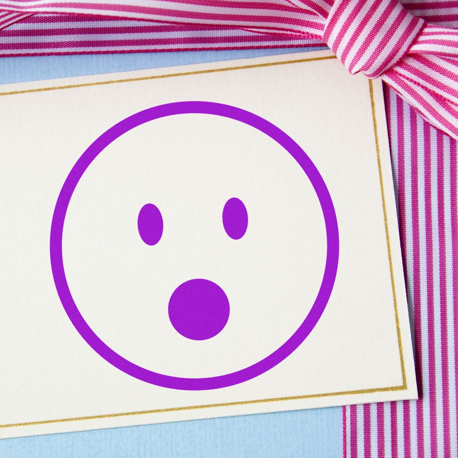 Self-Inking Gasping Gary Pictogram Stamp on a card with a purple surprised face, bordered in gold, next to a pink and white striped ribbon. Perfect for adding fun expressions to your projects.