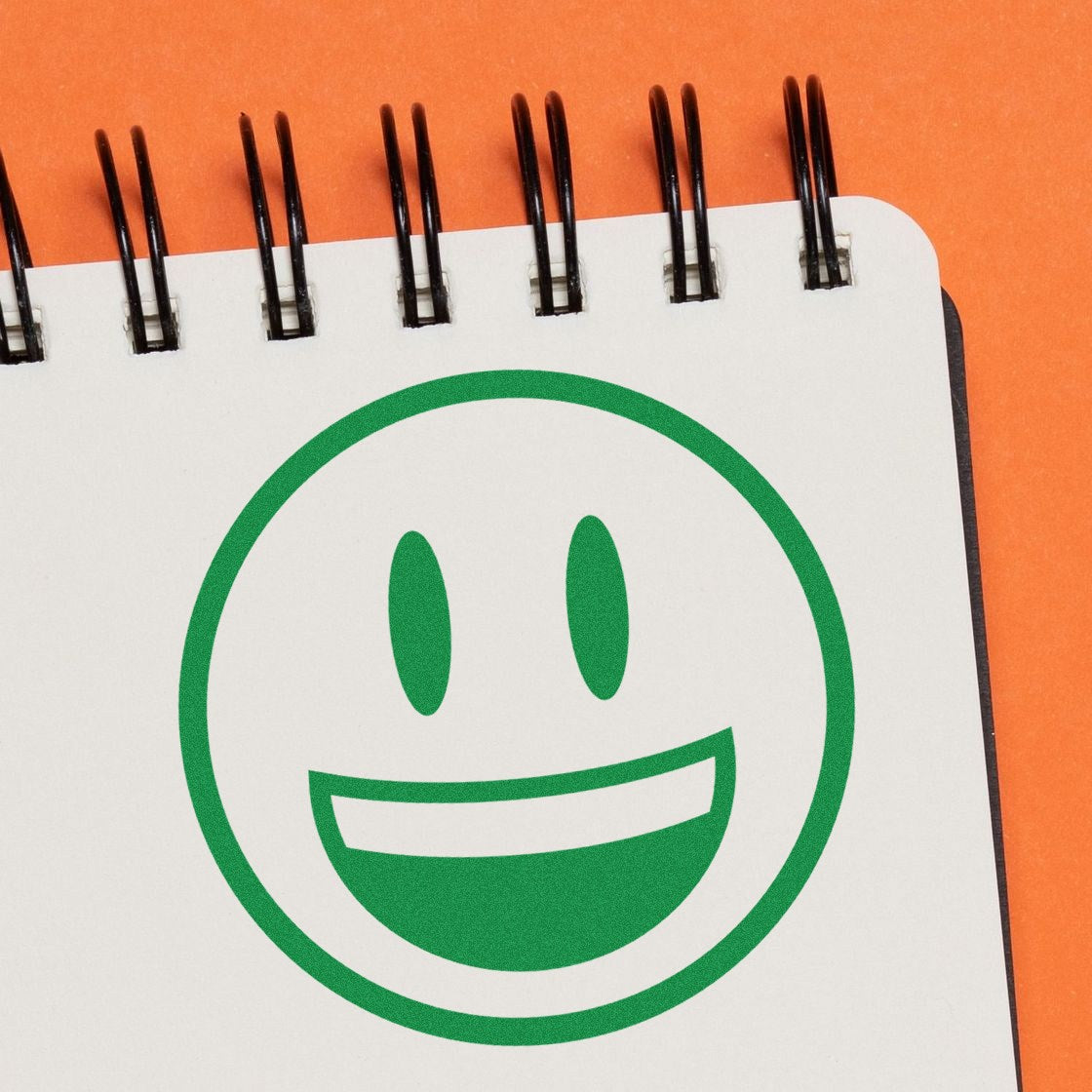 Self-Inking Grinning Delight Emoji Stamp on orange background, featuring a green smiling face on a notepad. Perfect for adding fun and personality to your stationery collection.