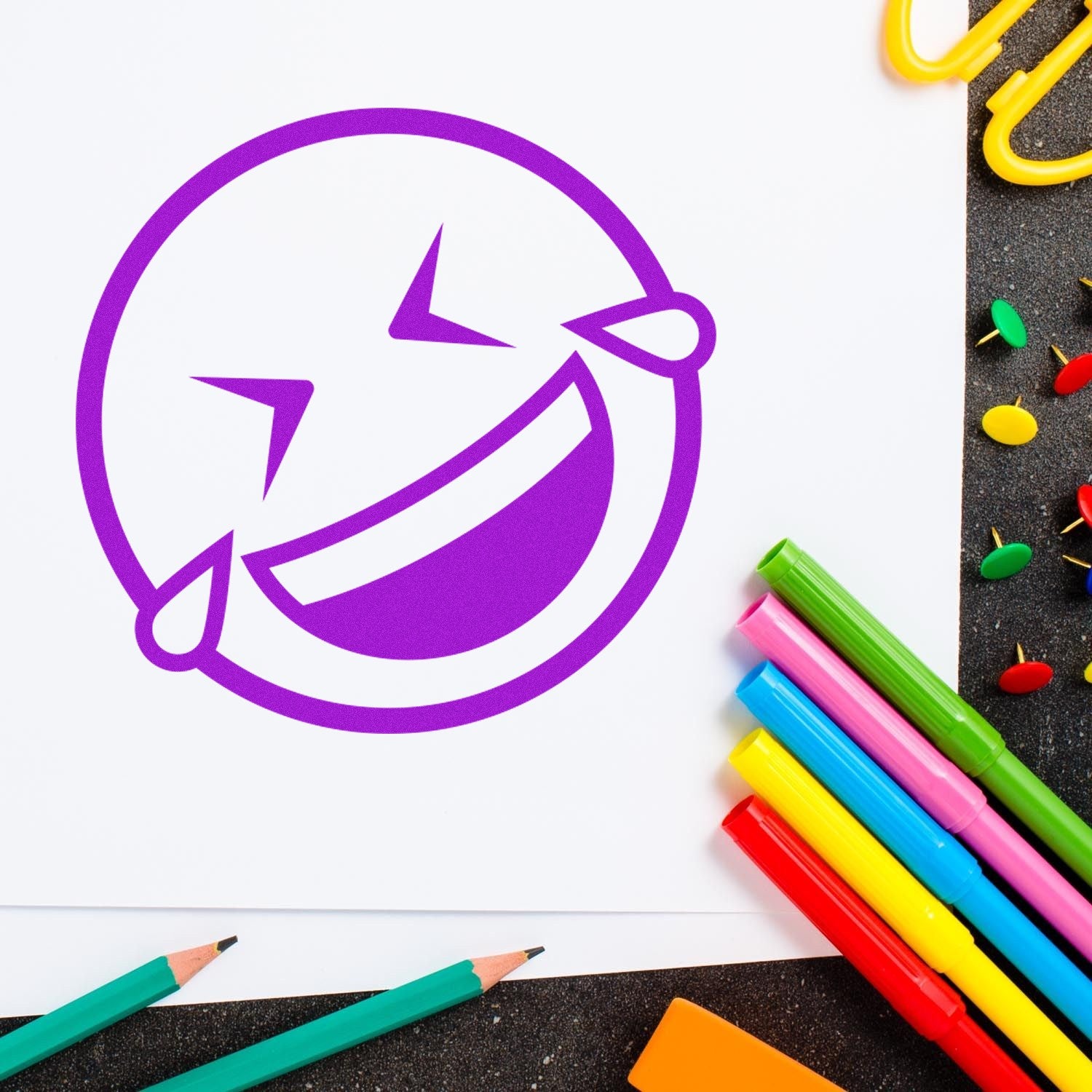 Wood Handle Laugh Riot Smiley Rubber Stamp imprint on paper, featuring a purple laughing emoji. Surrounded by colorful pens, pencils, and push pins on a dark surface.