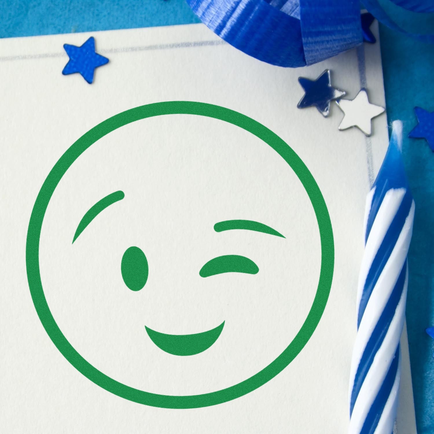 Self-Inking Winky Face Pictogram Stamp in green ink on paper, surrounded by blue stars and a striped candle, perfect for adding a playful touch to crafts and stationery.