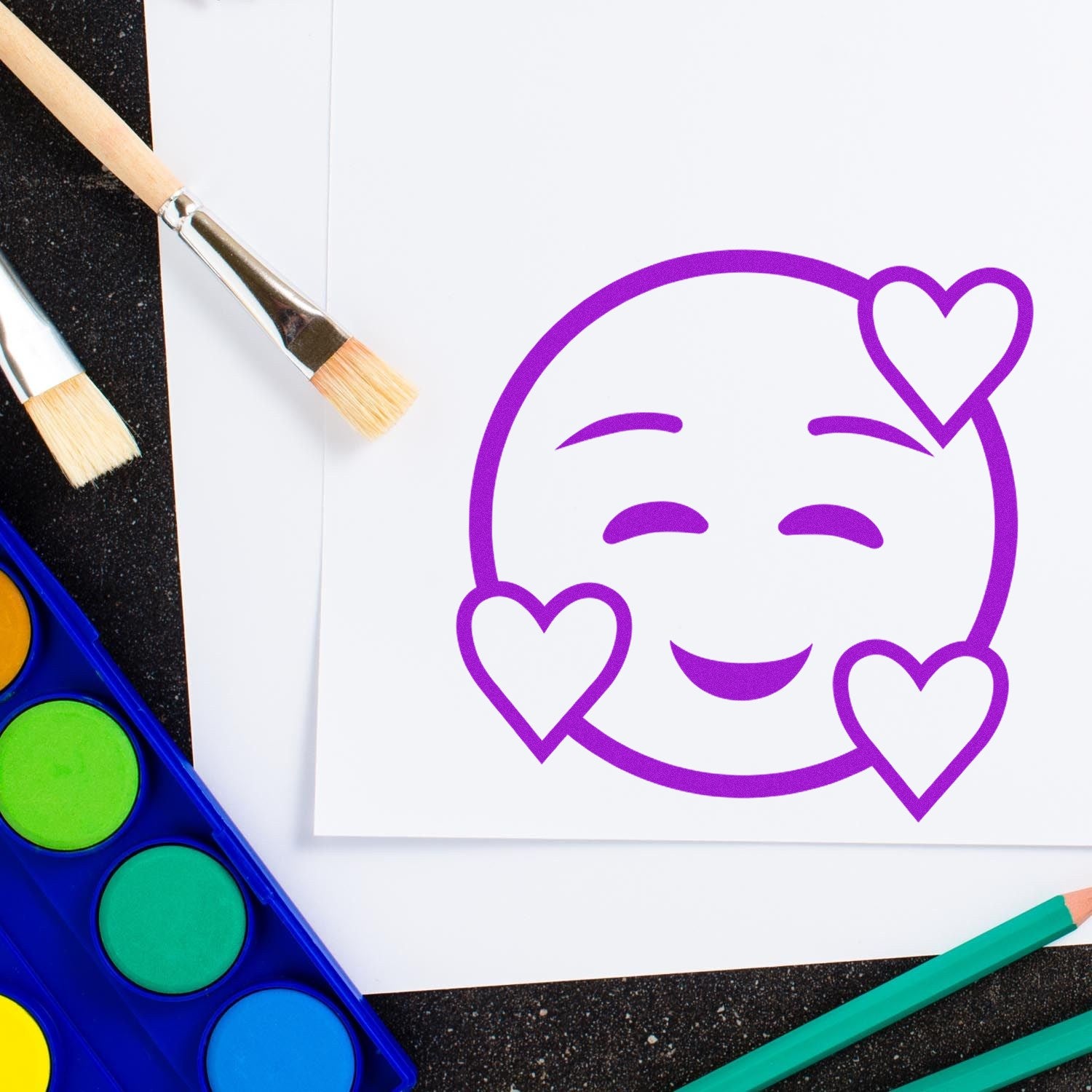 Wood Handle Love-Struck Emotes Rubber Stamp featuring a smiling face with heart eyes, shown on white paper. Includes wooden handle and vibrant purple ink. Perfect for crafts and decoration.