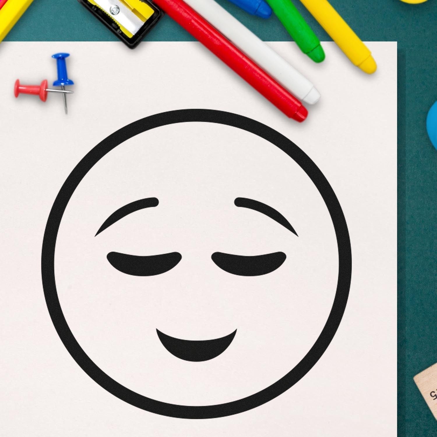 Self-Inking Contentment Facial Expressions Stamp on paper, surrounded by colorful pens and push pins, showcasing a serene smiley face design. Perfect for adding a touch of joy to any project.